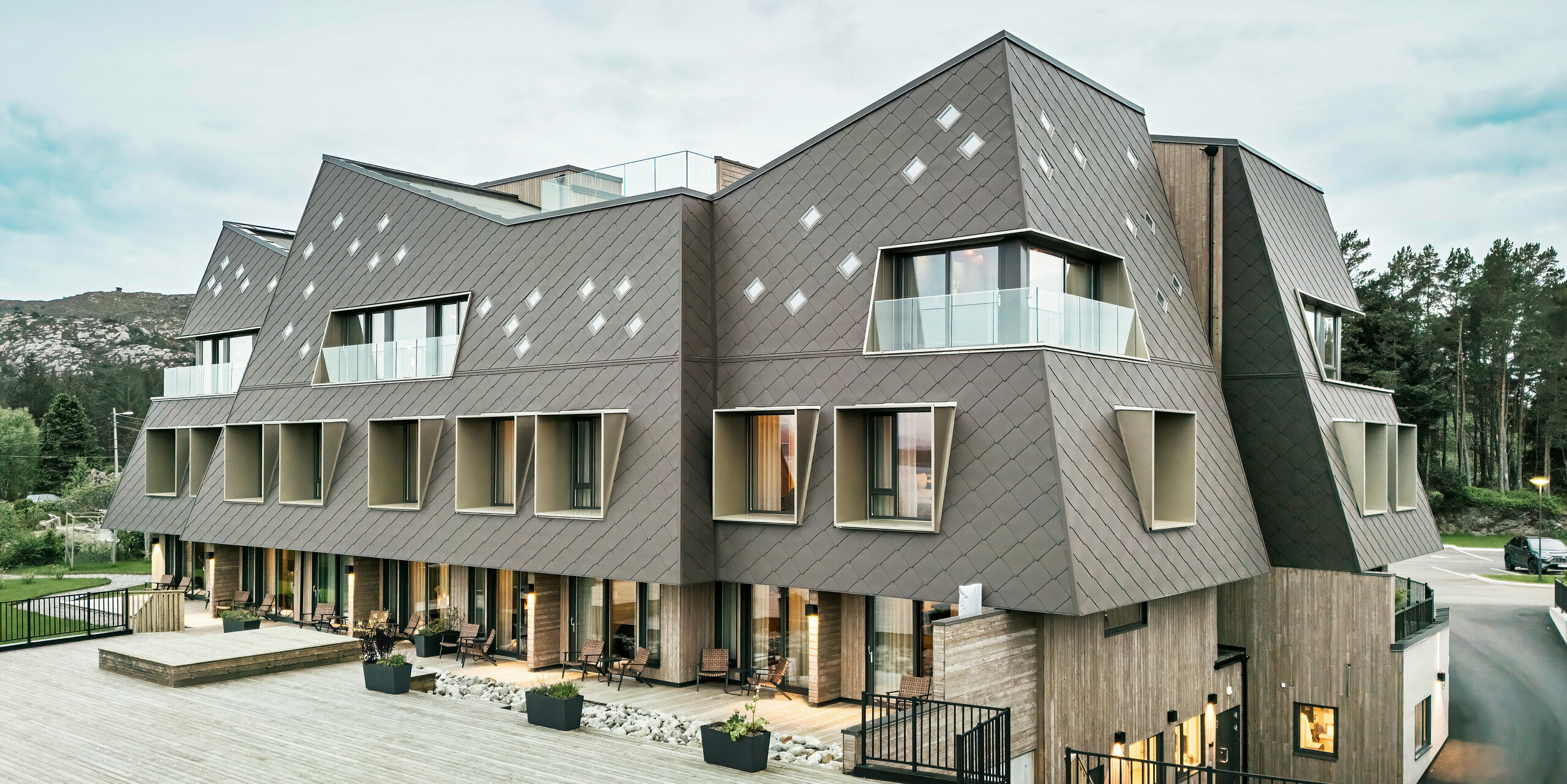 Vue de la terrasse de l'hôtel Beckerwyc House à Bekkjarvik, Norvège. Le bâtiment moderne est décoré d'une façade en aluminium frappante composée de losanges PREFA 44 × 44 en brun noisette. L'architecture futuriste avec des surfaces de toit en pente et des éléments de fenêtre proéminents est complétée par des espaces extérieurs spacieux. La couleur naturelle de la façade s'intègre harmonieusement dans le paysage environnant et souligne le design contemporain du bâtiment.