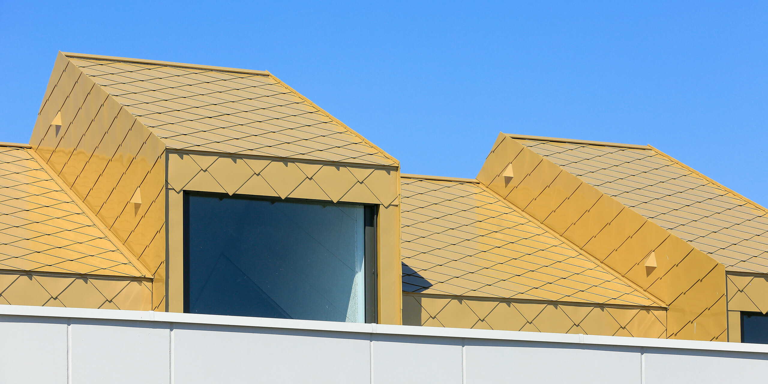 Gros plan sur une fenêtre de toit du centre communautaire et culturel de Fessenheim en France. La fenêtre est recouverte de losanges de toiture et de façade PREFA 44 × 44 en Sparkling Gold.