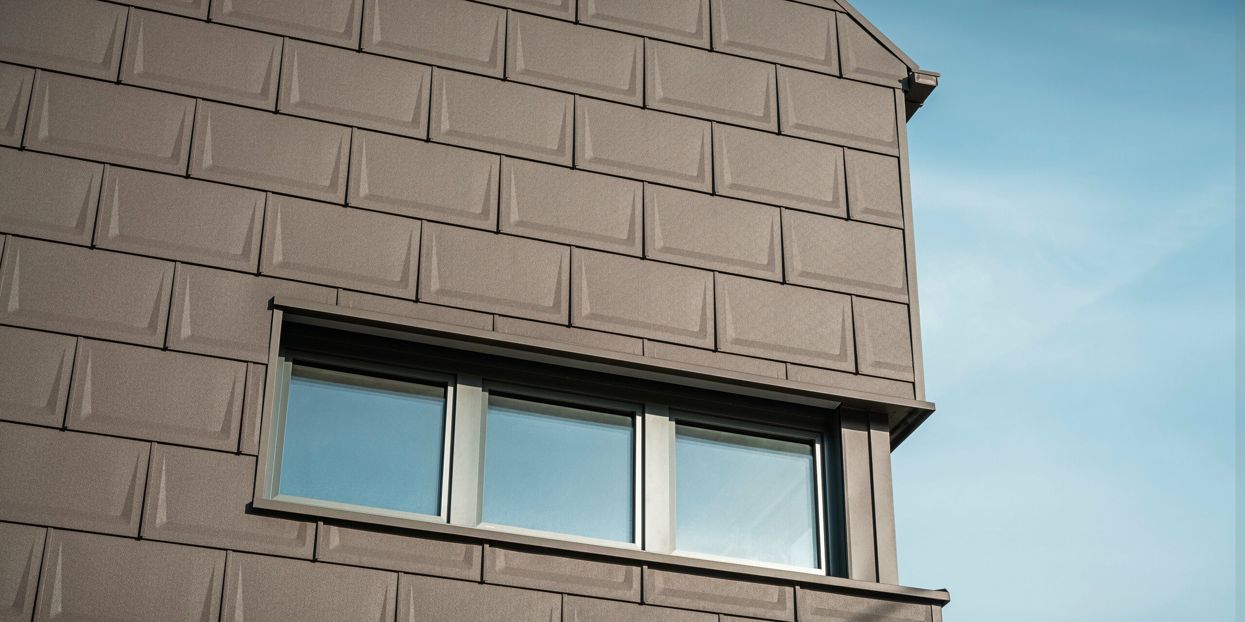 Nahaufnahme der Fassade eines Hauses in Neukirchen, Österreich, gestaltet mit der innovativen PREFA Dachplatte R.16 in braun P.10, die als Sonderbaulösung für die Dach- und Fassadengestaltung des Obergeschosses eingesetzt wird. Diese einzigartige Anwendung sorgt für ein harmonisches und ästhetisches Erscheinungsbild und unterstreicht die Vielseitigkeit der PREFA Aluminiumprodukte. Die einheitliche Farbgebung und das durchdachte Gestaltungskonzept des Gebäudes unterstreichen die vielseitigen Gestaltungsmöglichkeiten von PREFA.