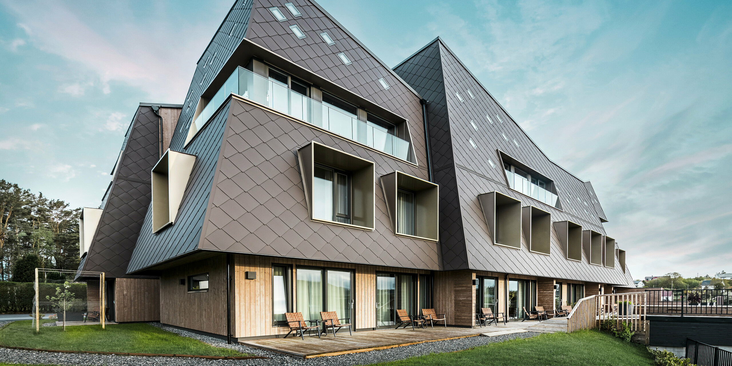 Vue plongeante sur le boutique-hôtel Beckerwyc House à Bekkjarvik, en Norvège, avec une façade saisissante composée de losanges de façade PREFA 44 × 44 en brun noisette. L'architecture inhabituelle du bâtiment, avec ses éléments de façade inclinés et ses fenêtres saillantes, crée une apparence futuriste qui s'intègre harmonieusement dans l'environnement naturel. Le design moderne et l'utilisation de matériaux de haute qualité soulignent le design exclusif de l'hôtel.