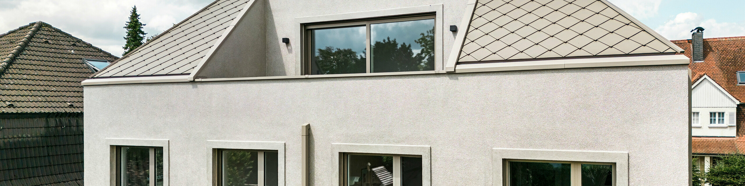 Vue de l'étage supérieur d'une maison familiale moderne avec un toit en croupe et une toiture en aluminium couleur bronze de PREFA. Le toit est recouvert d'élégants losanges en aluminium qui soulignent le design contemporain et élégant de la maison. L'étage supérieur dispose de grandes fenêtres laissant entrer beaucoup de lumière naturelle. La façade de la maison présente un design simple et moderne, ce qui confère au bâtiment un aspect élégant et harmonieux. En arrière-plan, vous pouvez voir d'autres maisons du quartier, qui soulignent l'aspect général harmonieux du quartier résidentiel.