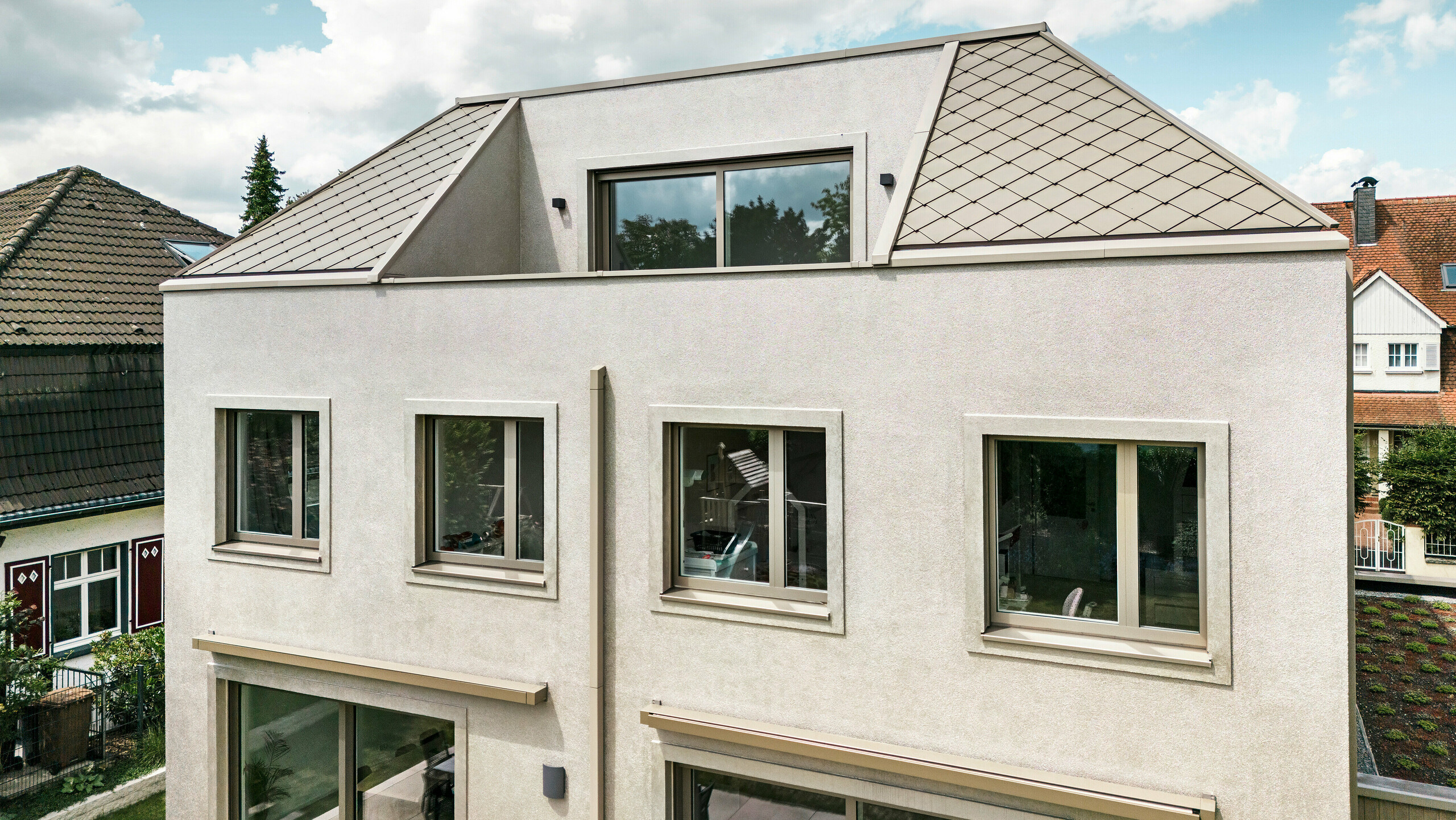 Vue de l'étage supérieur d'une maison familiale moderne avec un toit en croupe et une toiture en aluminium couleur bronze de PREFA. Le toit est recouvert d'élégants losanges en aluminium qui soulignent le design contemporain et élégant de la maison. L'étage supérieur dispose de grandes fenêtres laissant entrer beaucoup de lumière naturelle. La façade de la maison présente un design simple et moderne, ce qui confère au bâtiment un aspect élégant et harmonieux. En arrière-plan, vous pouvez voir d'autres maisons du quartier, qui soulignent l'aspect général harmonieux du quartier résidentiel.