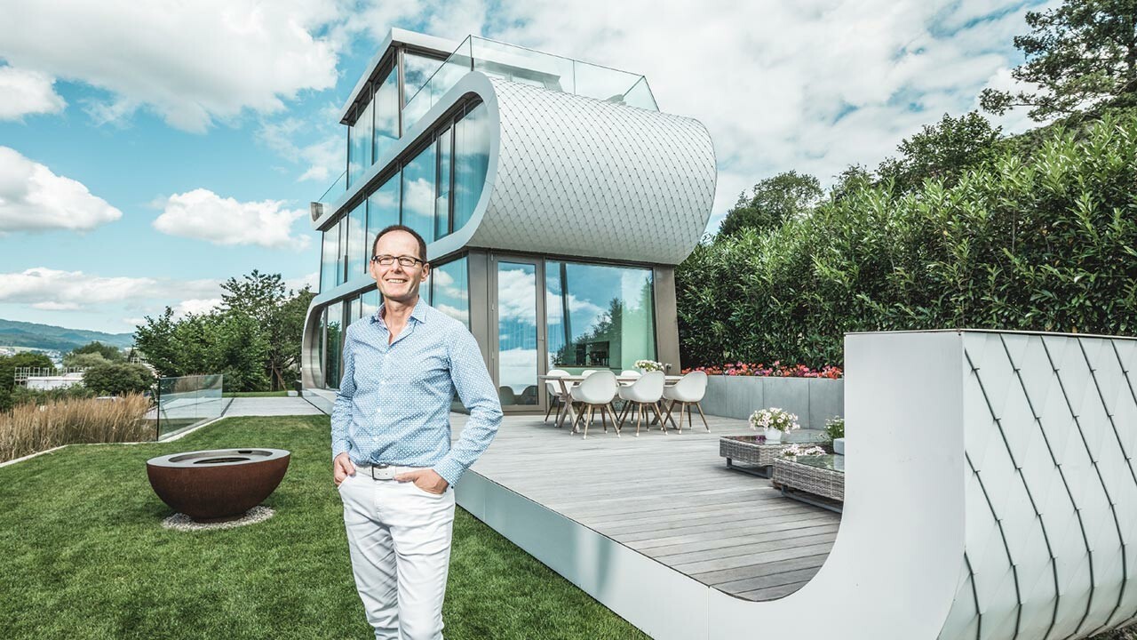 Der Architekt Stefan Camenzind steht in weißer Hose und hellblauem Hemd vor dem Flexhouse am Zürichsee. Die gebogene Fassade wurde mit reinweißen Alu-Schindeln von PREFA verkleidet. 