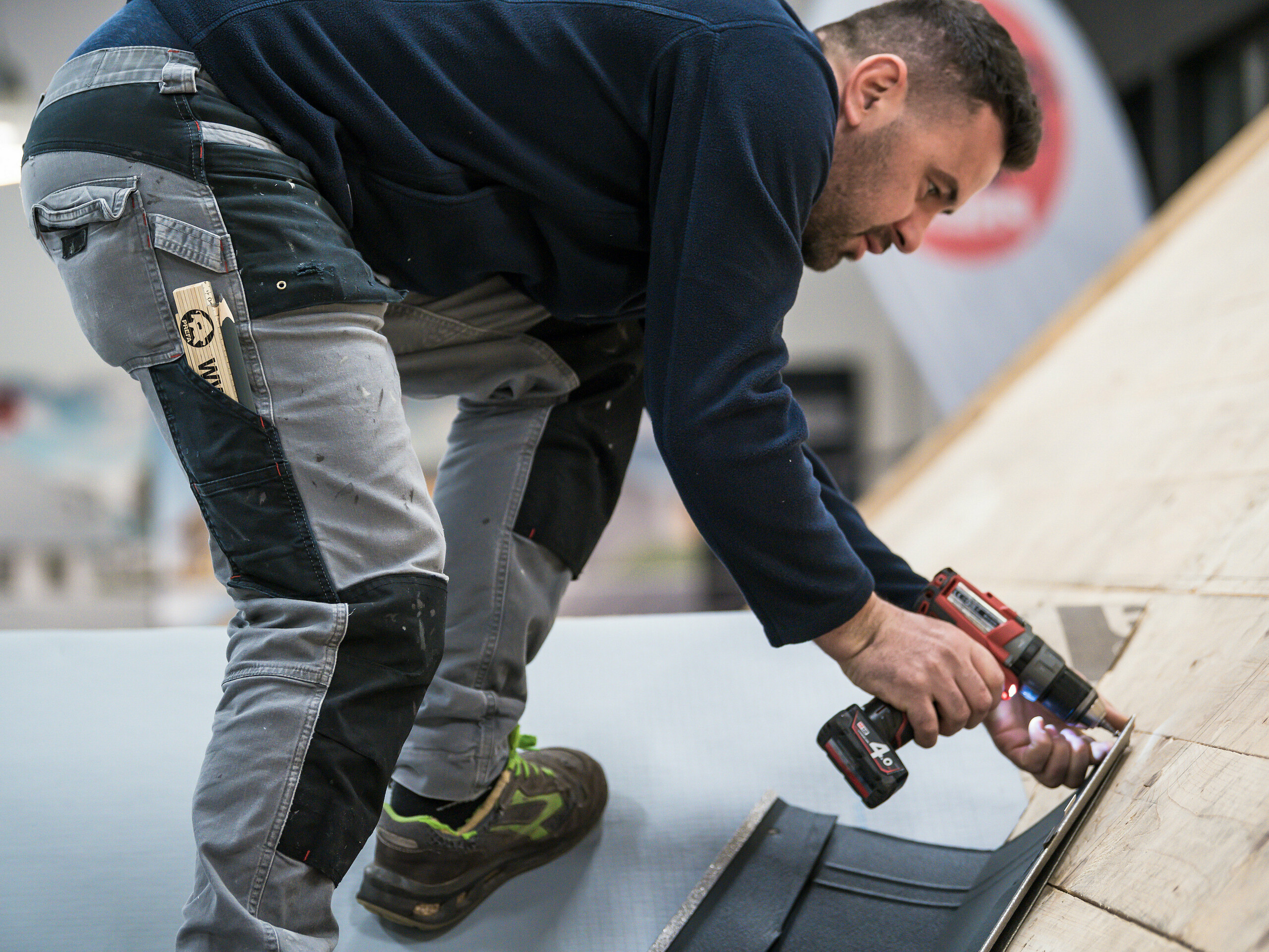 Zu sehen ist ein Handwerker in der PREFA Academy, der gerade mit einer Bohrmaschine eine PREFA Dachplatte auf einem Dach montiert.