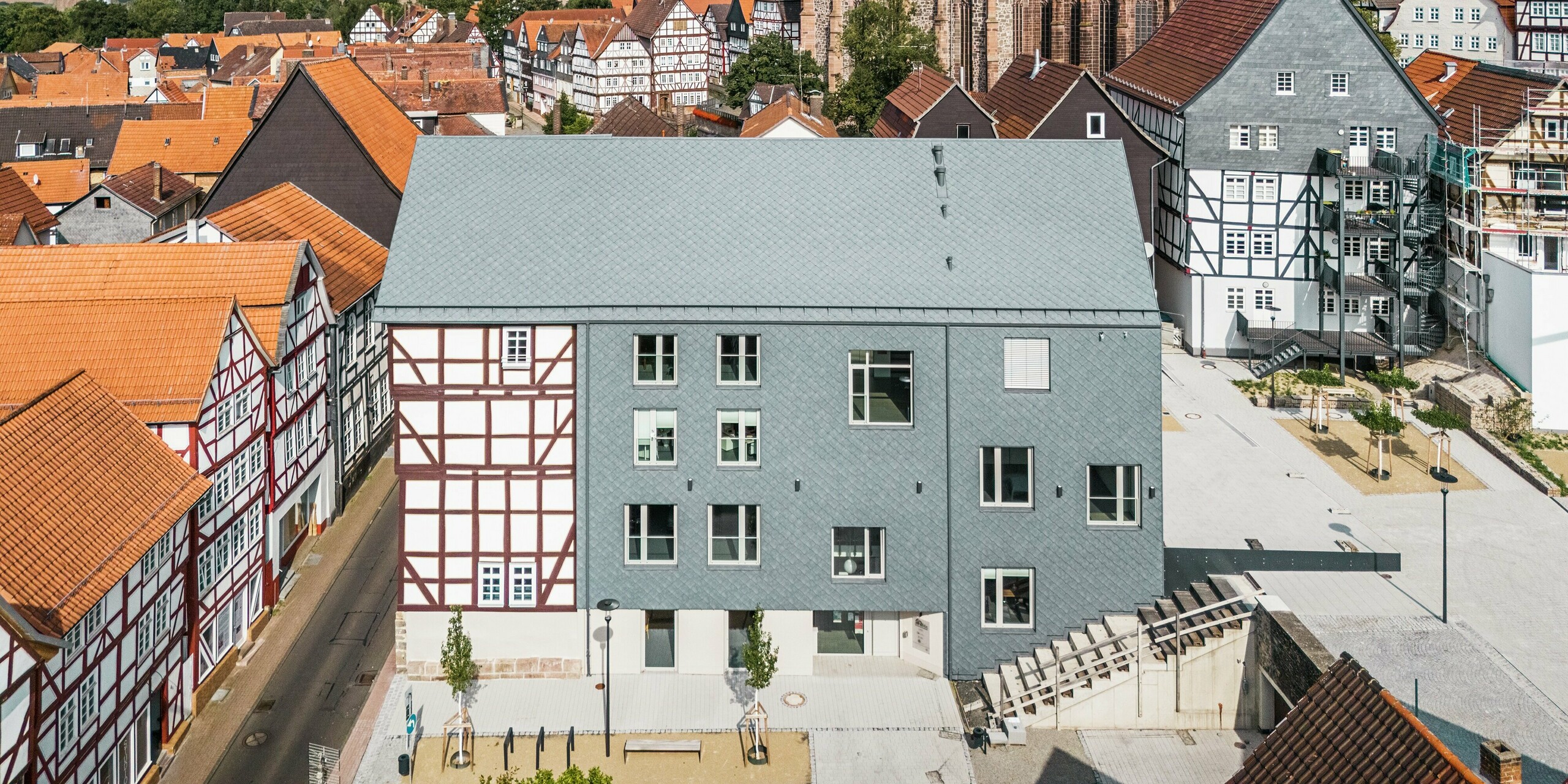 Drohnenaufnahme des Volkshauses in Homberg, DE, mit PREFA Dach- und Wandrauten 29 × 29 in P.10 Steingrau. Das modern sanierte Gebäude harmoniert dank der langlebigen und witterungsbeständigen Aluminiumbekleidung perfekt mit den umliegenden Fachwerkhäusern und der historischen Kirche.