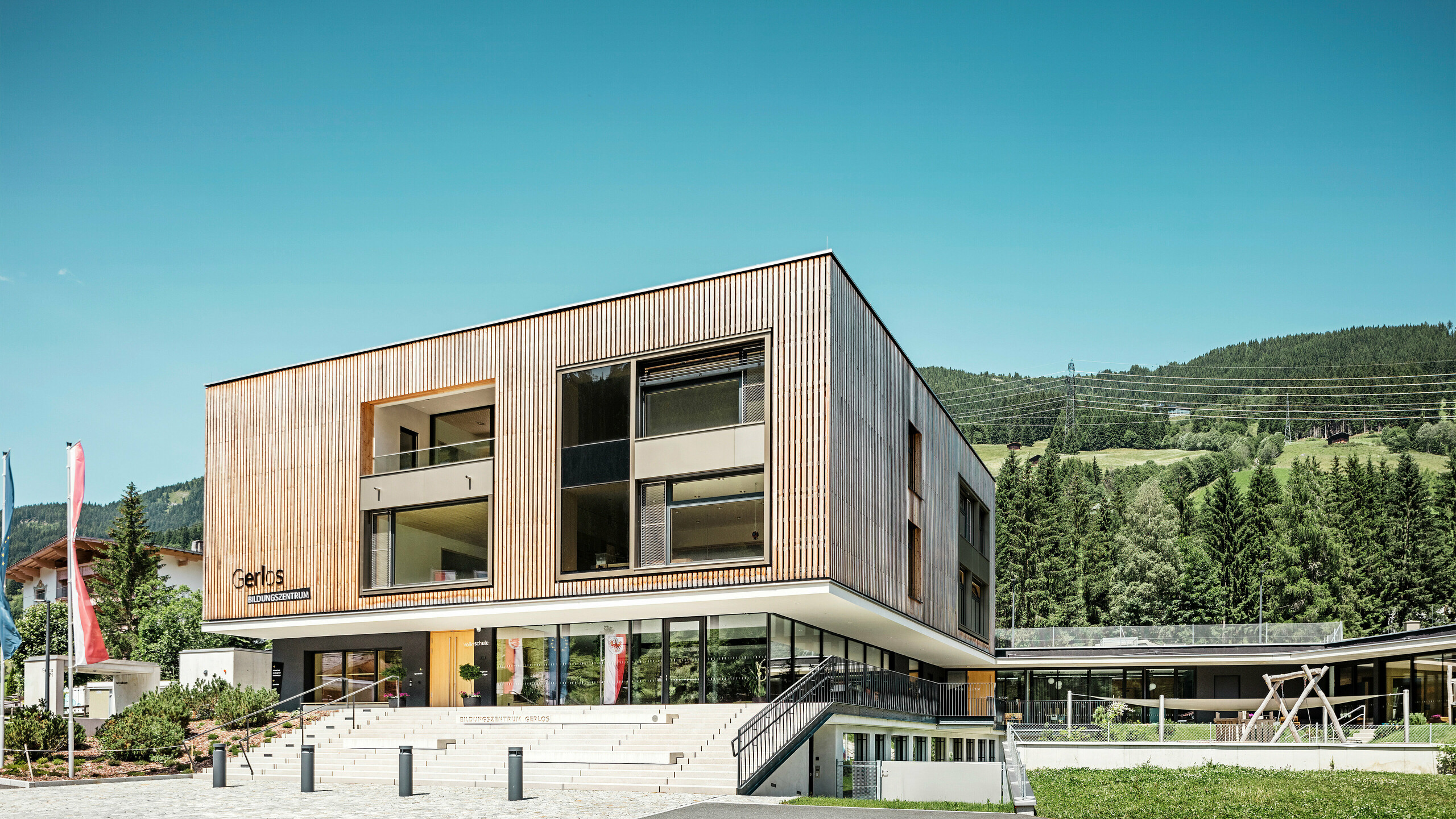 Vue de face du centre de formation moderne avec une façade en bois.  Le bâtiment s'élève sur deux étages avec de grandes baies vitrées et un toit plat qui s'intègre parfaitement dans le paysage montagneux environnant avec ses collines densément boisées. Au premier plan, on peut voir des espaces verts bien entretenus, une voie d'accès en gradins et des drapeaux publicitaires. Le design transmet une combinaison de fonctionnalité et d'architecture esthétique en harmonie avec la nature.