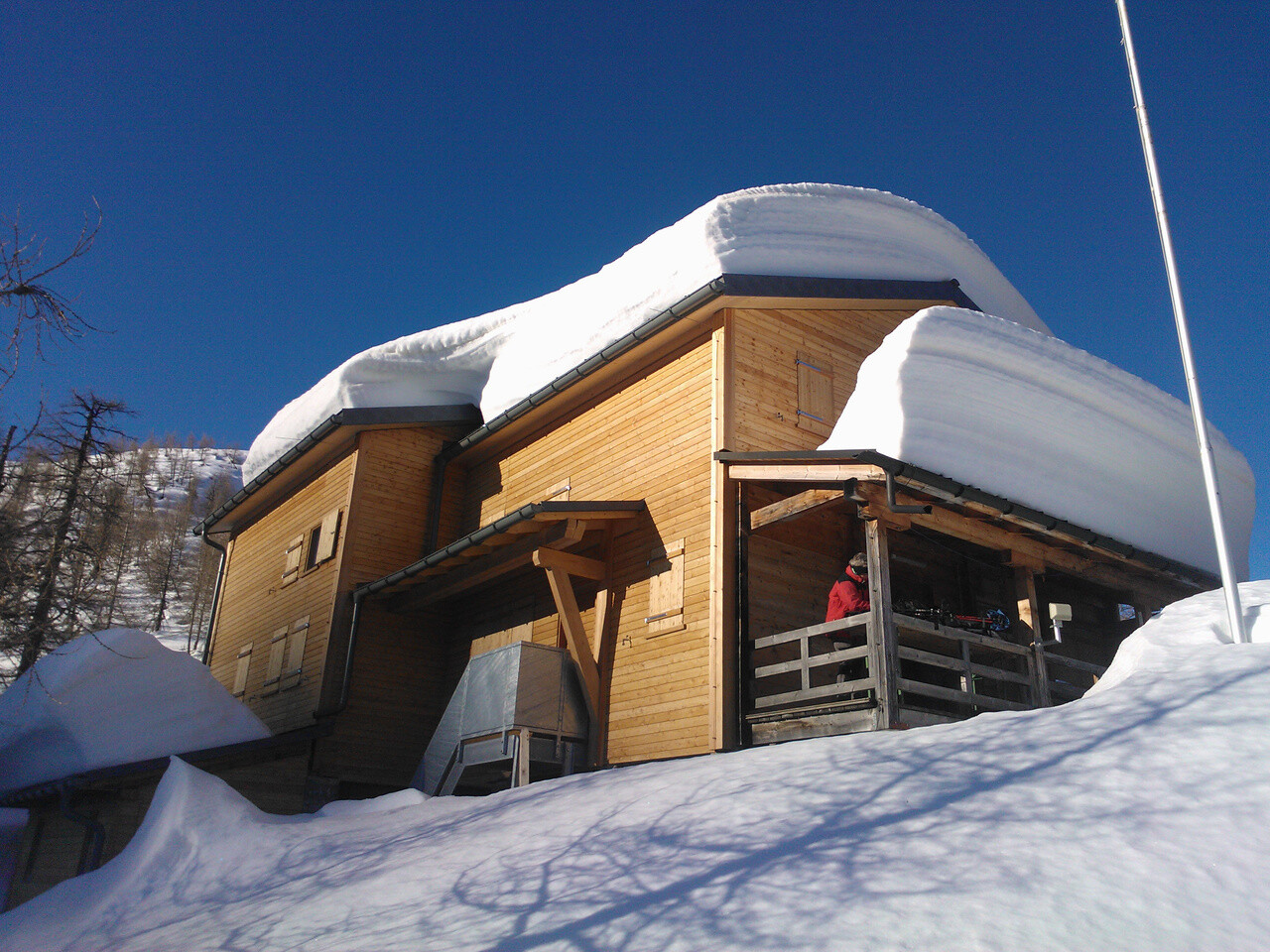 Das Bild zeigt die Hütte Capanna Buffaloraim im Winter, die von dicken Schneewechten am Dach bedeckt ist. Das Bild weist auf die Gefahren durch abrutschende Schneelawinen hin. Das Dach der Berghütte ist mit robusten PREFA Dachpaneelen FX.12-gedeckt, welche besonders für extreme Wetterbedingungen geeignet sind. Trotz der massiven Schneelast bleibt die Dachkonstruktion stabil und sicher. Dies zeigt die hohe Belastbarkeit und Qualität der PREFA Produkte. Die Berghütte selbst besteht aus Holz und fügt sich harmonisch in die winterliche Landschaft ein.