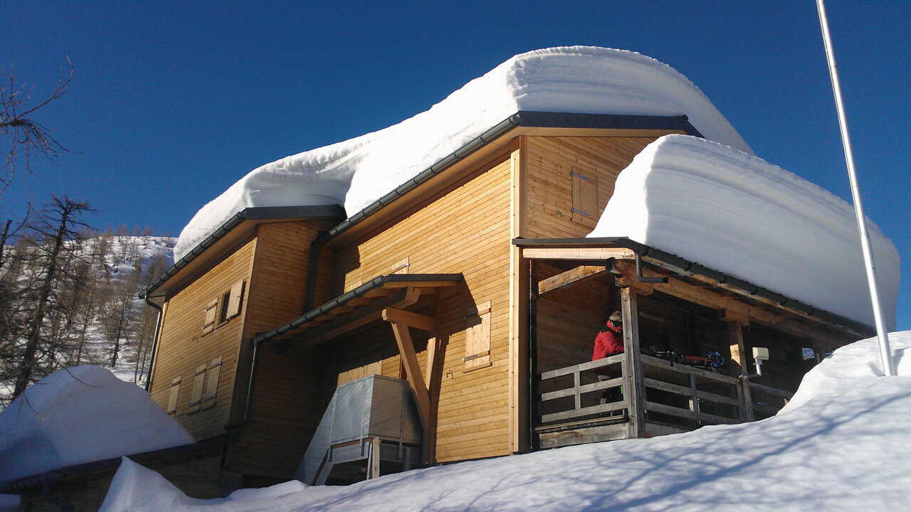 Das Bild zeigt die Hütte Capanna Buffaloraim im Winter, die von dicken Schneewechten am Dach bedeckt ist. Das Bild weist auf die Gefahren durch abrutschende Schneelawinen hin. Das Dach der Berghütte ist mit robusten PREFA Dachpaneelen FX.12-gedeckt, welche besonders für extreme Wetterbedingungen geeignet sind. Trotz der massiven Schneelast bleibt die Dachkonstruktion stabil und sicher. Dies zeigt die hohe Belastbarkeit und Qualität der PREFA Produkte. Die Berghütte selbst besteht aus Holz und fügt sich harmonisch in die winterliche Landschaft ein.