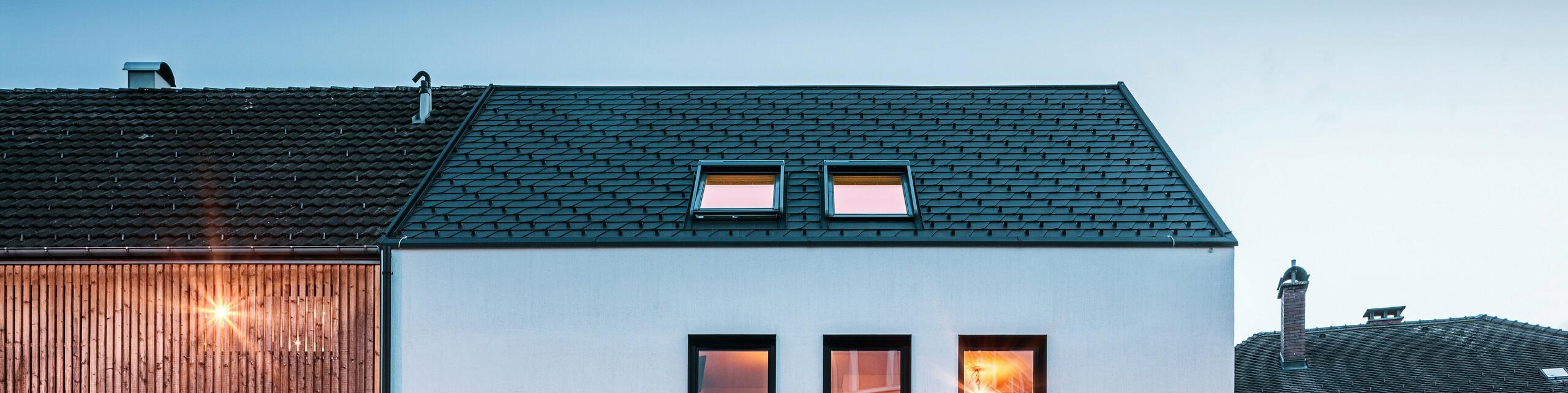 Moderne Mehrfamilienhausfassade mit PREFA Dachschindeln DS19 und Prefalz in Schwarz am Bodensee in Österreich. Das Gebäude hat eine klare Zweiteilung mit einem hölzernen Anbau auf der linken Seite und einem verputzten Hauptteil auf der rechten Seite. Das Dach zeigt die charakteristische dunkle Schindeldeckung und es gibt mehrere rechteckige Fenster, die im warmen Abendlicht spiegeln.