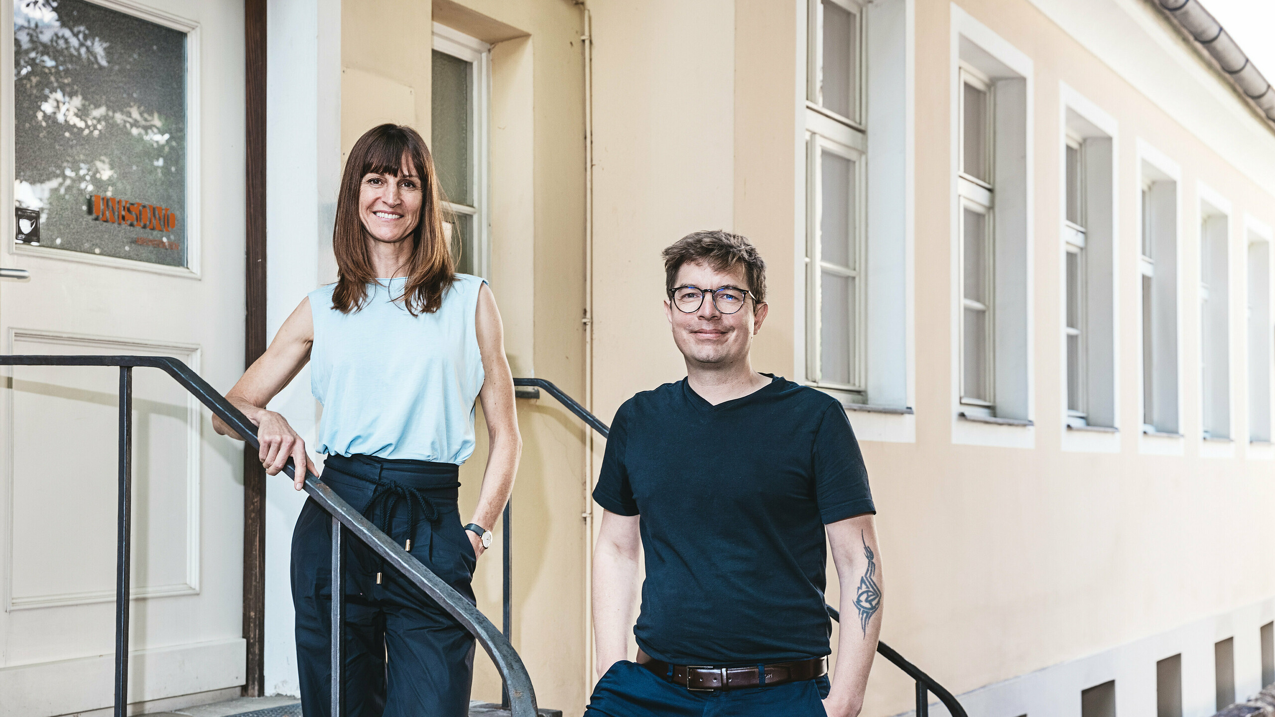 Les principaux architectes de l'agence UNISONO ARCHITEKTEN d'Innsbruck se tiennent sur un escalier en pierre devant un bâtiment peint en beige avec des cadres de fenêtres blancs. La femme porte un chemisier bleu sans manches et un pantalon noir, l'homme un T-shirt noir et un pantalon bleu avec une ceinture marron. Ils sourient aimablement à l'appareil photo, suggérant une atmosphère ouverte et accueillante.