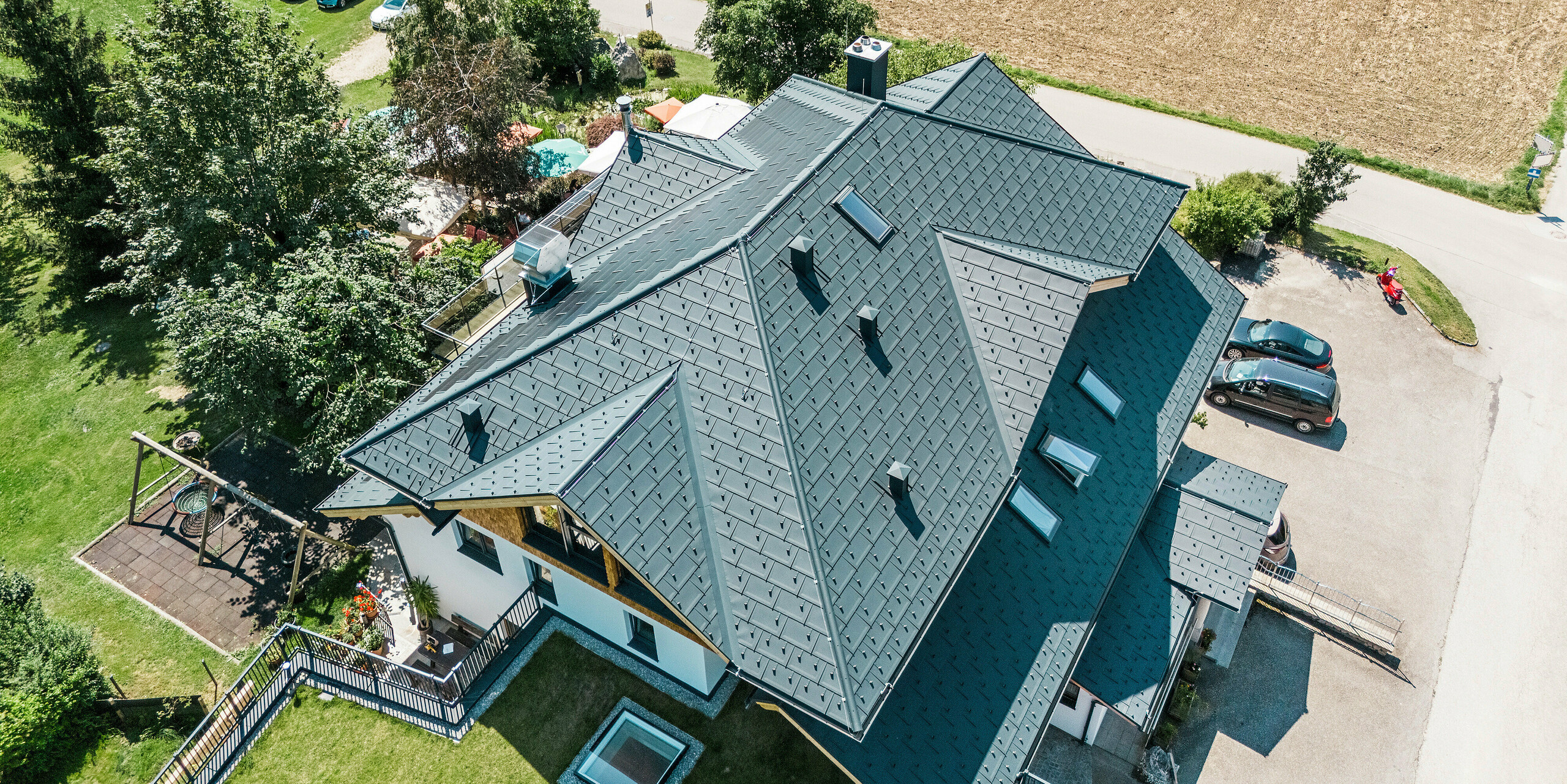 Blick von oben auf das Dach der Zenz'n Stub'n in Schörfling am Attersee, eingedeckt mit PREFA Dachplatten R.16 und Solardachplatten in der hochwertigen Beschichtung P.10 Anthrazit. Das innovative Aluminiumdach verbindet moderne Ästhetik mit nachhaltiger Solartechnik und fügt sich harmonisch in die ländliche Umgebung ein. Das langlebige PREFA Dachsystem bietet umfassenden Schutz und eine stilvolle Optik.