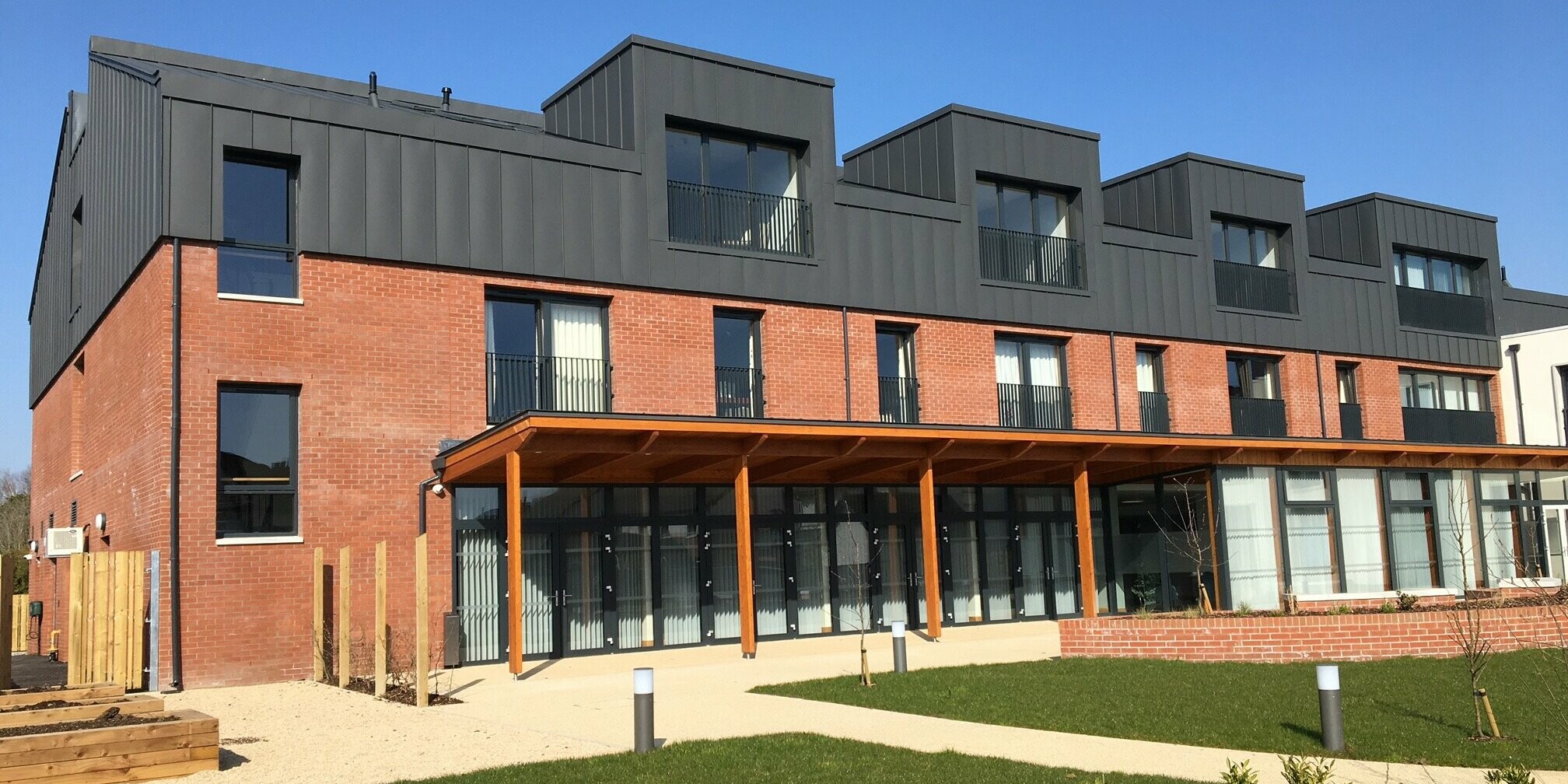 Rückansicht der Harberton Hall in Belfast mit großzügigem Gartenbereich. Die PREFALZ Aluminiumbekleidung in P.10 dunkelgrau und die klassischen Ziegelwände ergeben ein stilvolles Gesamtbild. Die innovative Stehfalztechnik unterstreicht die Funktionalität und Ästhetik der Dachlandschaft mit mehreren Flachdachgauben. 