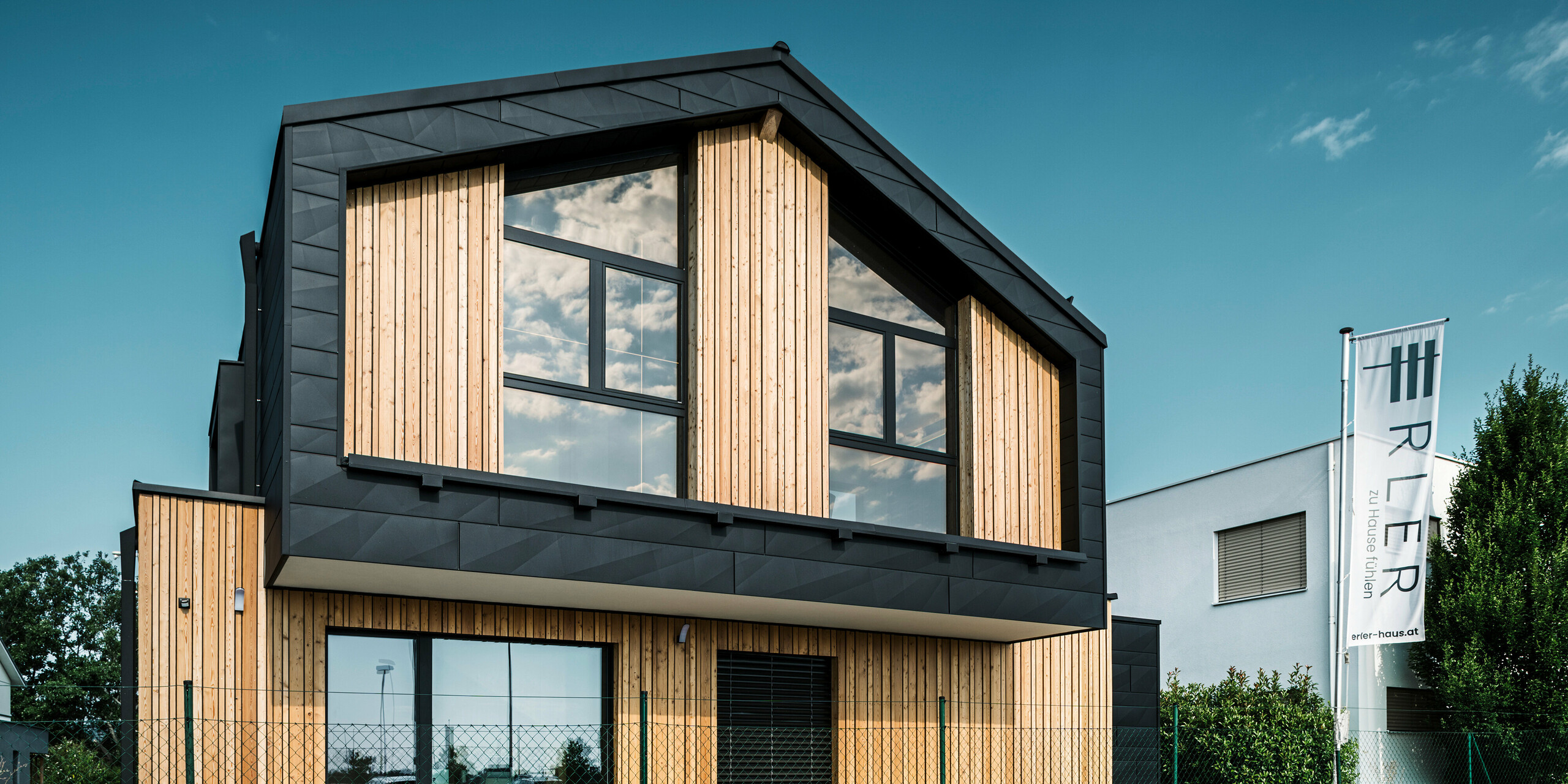 Stirnseite des Musterhauses Autark im Fertighauszentrum Blaue Lagune, Vösendorf (AT). Die Fassade kombiniert vertikale Holzelemente mit den schwarzen PREFA Siding.X Paneelen aus Aluminium. Große Glasflächen am Giebel reflektieren den Himmel und verleihen dem Haus ein modernes, lichtdurchflutetes Erscheinungsbild. Das Design betont die Harmonie zwischen Holz, Glas und Aluminium für eine zeitgemäße und nachhaltige Architektur.