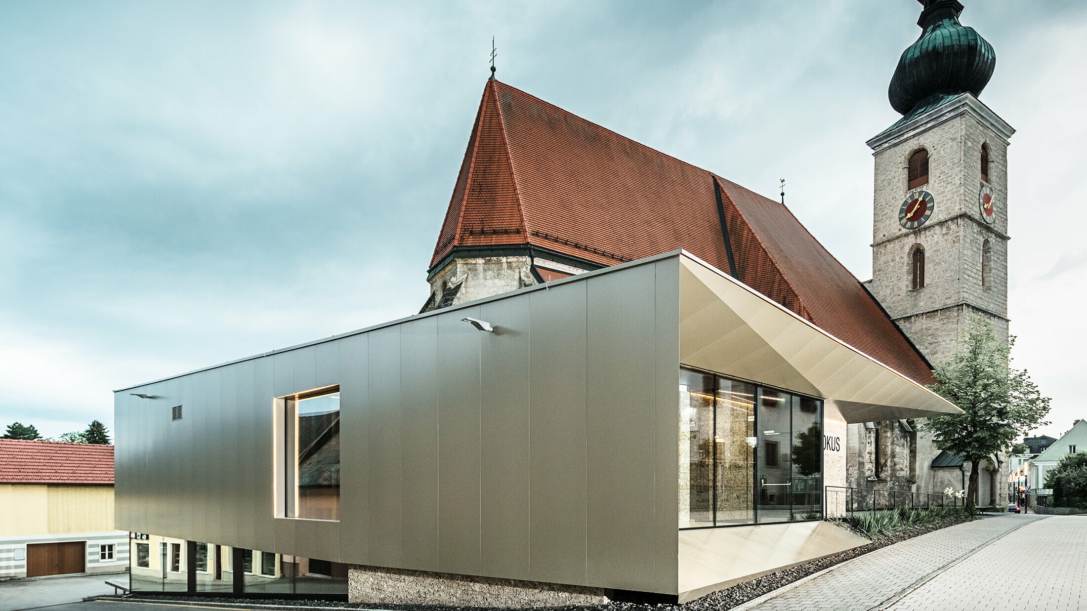 Die markante bronzefarbene Verbundplatten-Fassade des Pfarrzentrums in Sierning reckt sich um die kleine Kirche im Hintergrund