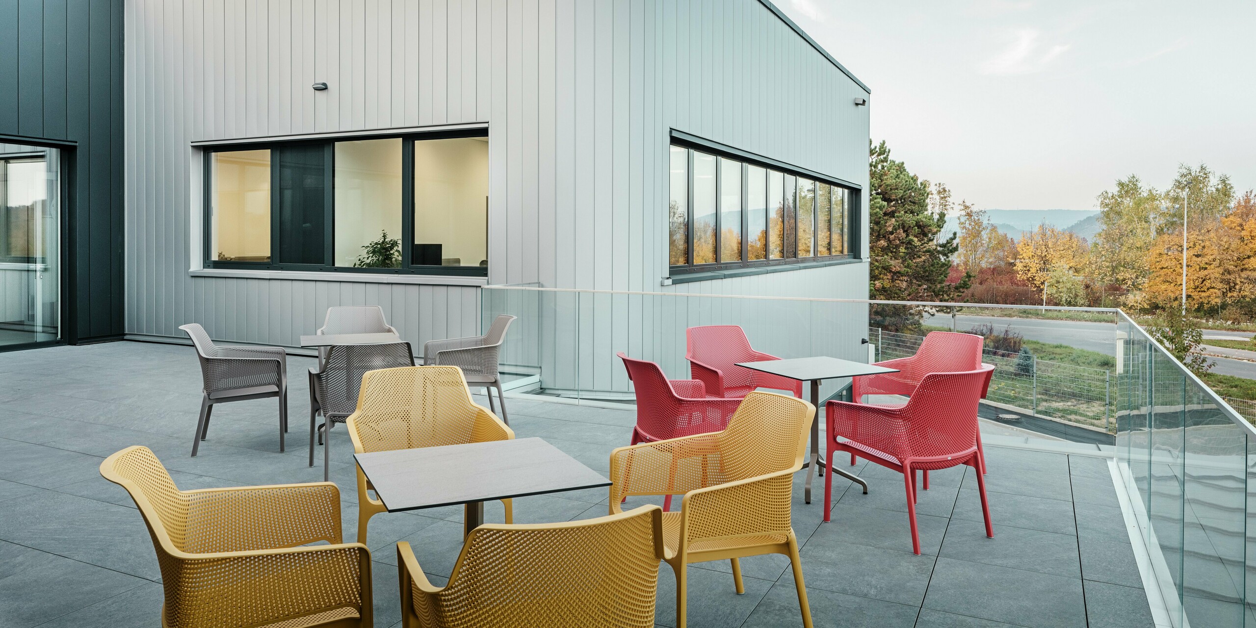 Die moderne Terrasse des neuen PREFA Standortes in Törökbálint bietet einen einladenden Außenbereich mit Blick auf die herbstliche Landschaft. Die Fassade ist mit PREFA Sidings in Silbermetallic und Anthrazit verkleidet, die sich durch ihre elegante Optik, hohe Witterungsbeständigkeit und Pflegeleichtigkeit auszeichnen. Die präzise Verarbeitung und die geradlinige, vertikale Gestaltung unterstreichen das zeitgemäße Design des Gebäudes. Die farbenfrohen Sitzmöbel im Vordergrund setzen farbige Akzente in der herbstlichen Kulisse.