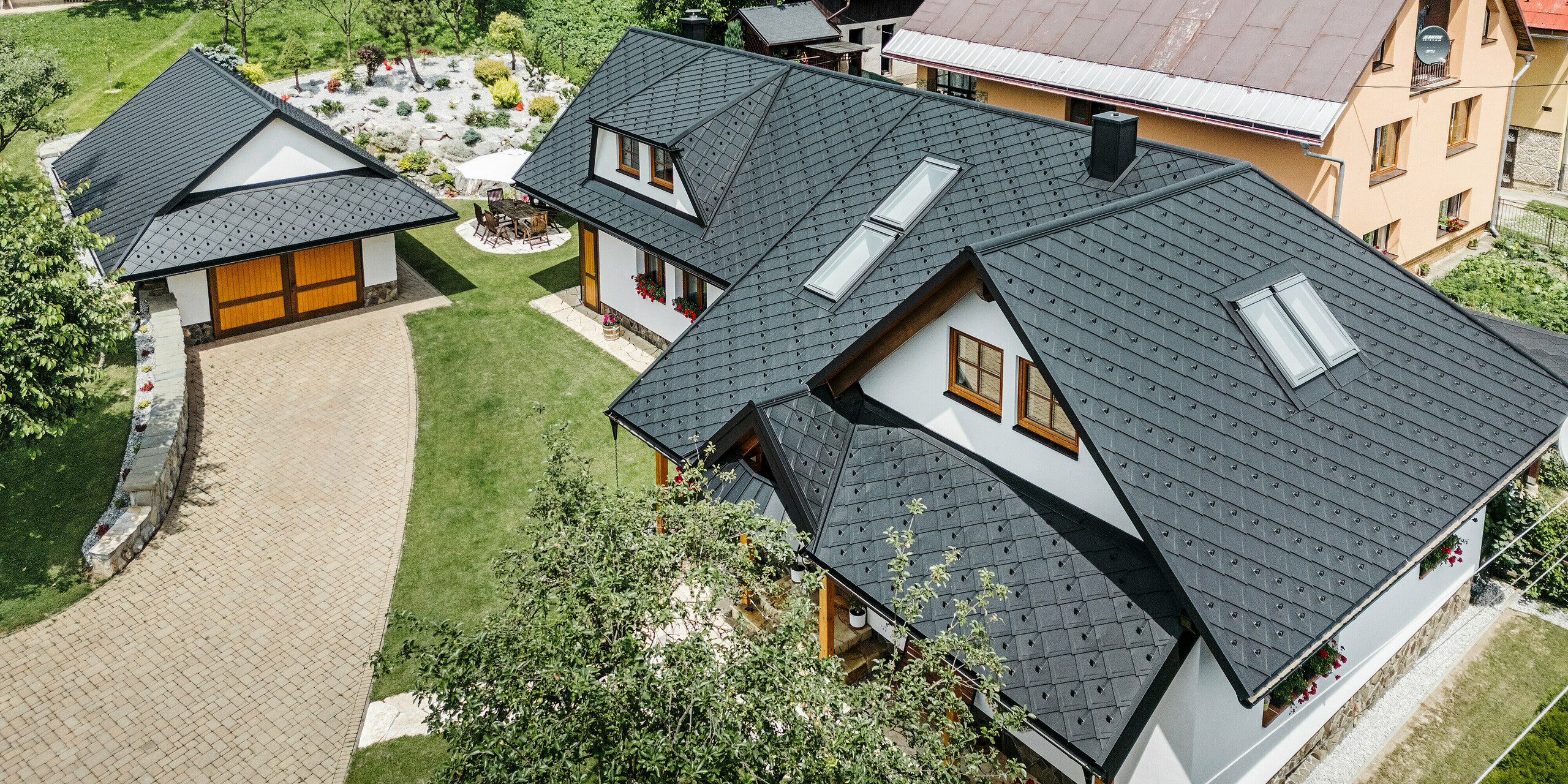 Vue aérienne d'une maison familiale à Stara Bystrica, en Slovaquie, avec un toit en losanges PREFA 29 × 29 en P.10 noir. Le toit, conçu dans un aspect de diamant uniforme, comprend des fenêtres de toit inclinées et des lucarnes trapézoïdales des deux côtés. Les décorations en bois sur les fenêtres et les compositions florales colorées confèrent à la maison un aspect invitant et harmonieux. Le jardin de devant bien entretenu et l'allée pavée complètent parfaitement l'ensemble.