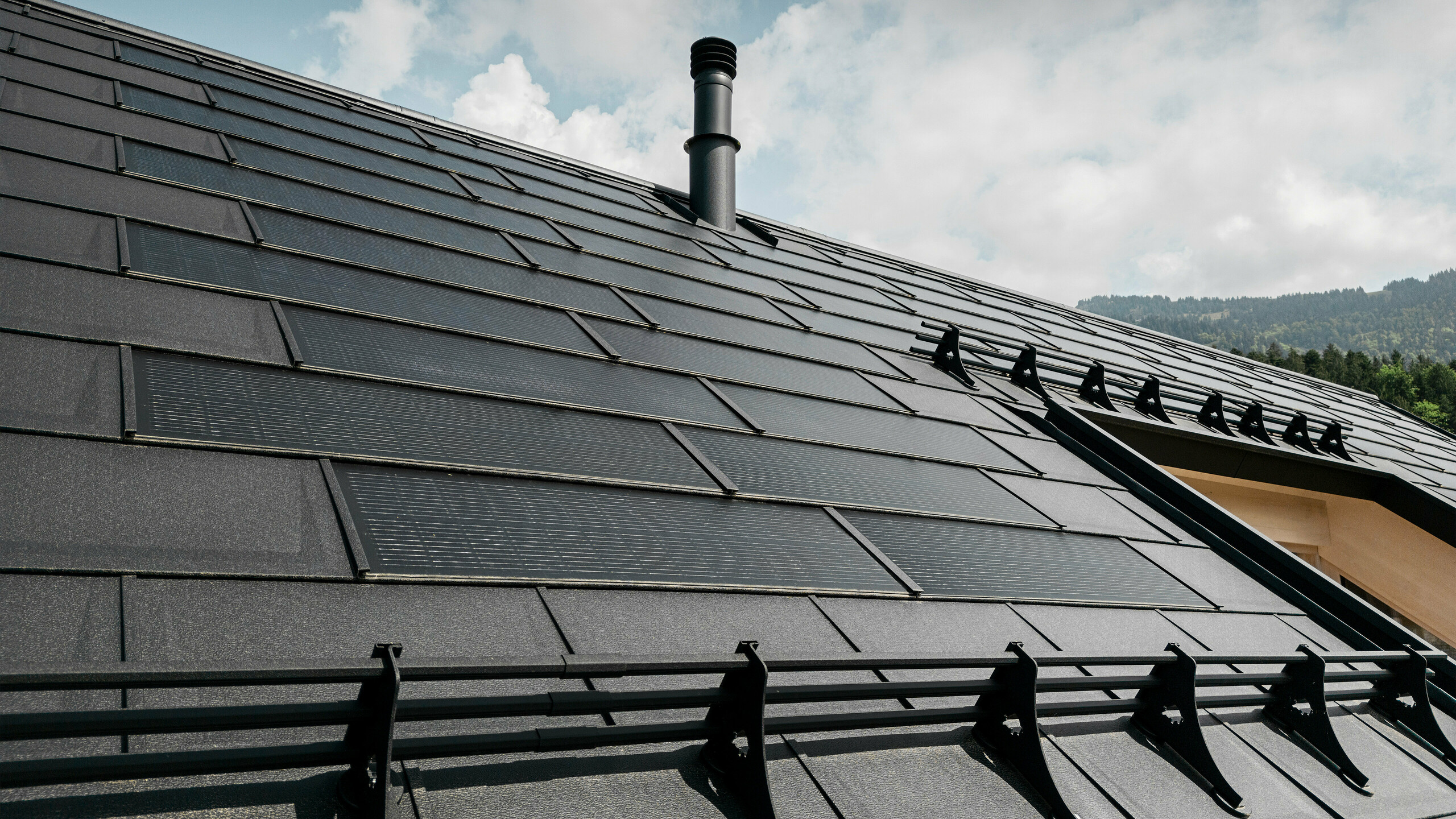 Gros plan sur la tuile solaire innovante PREFA, qui s'intègre parfaitement dans le toit noir composé de R.16 PREFA robustes. La combinaison d’une production d’énergie durable et d’une toiture en aluminium durable offre une solution de toiture écologique et esthétique qui s’intègre harmonieusement dans le paysage naturel. ​