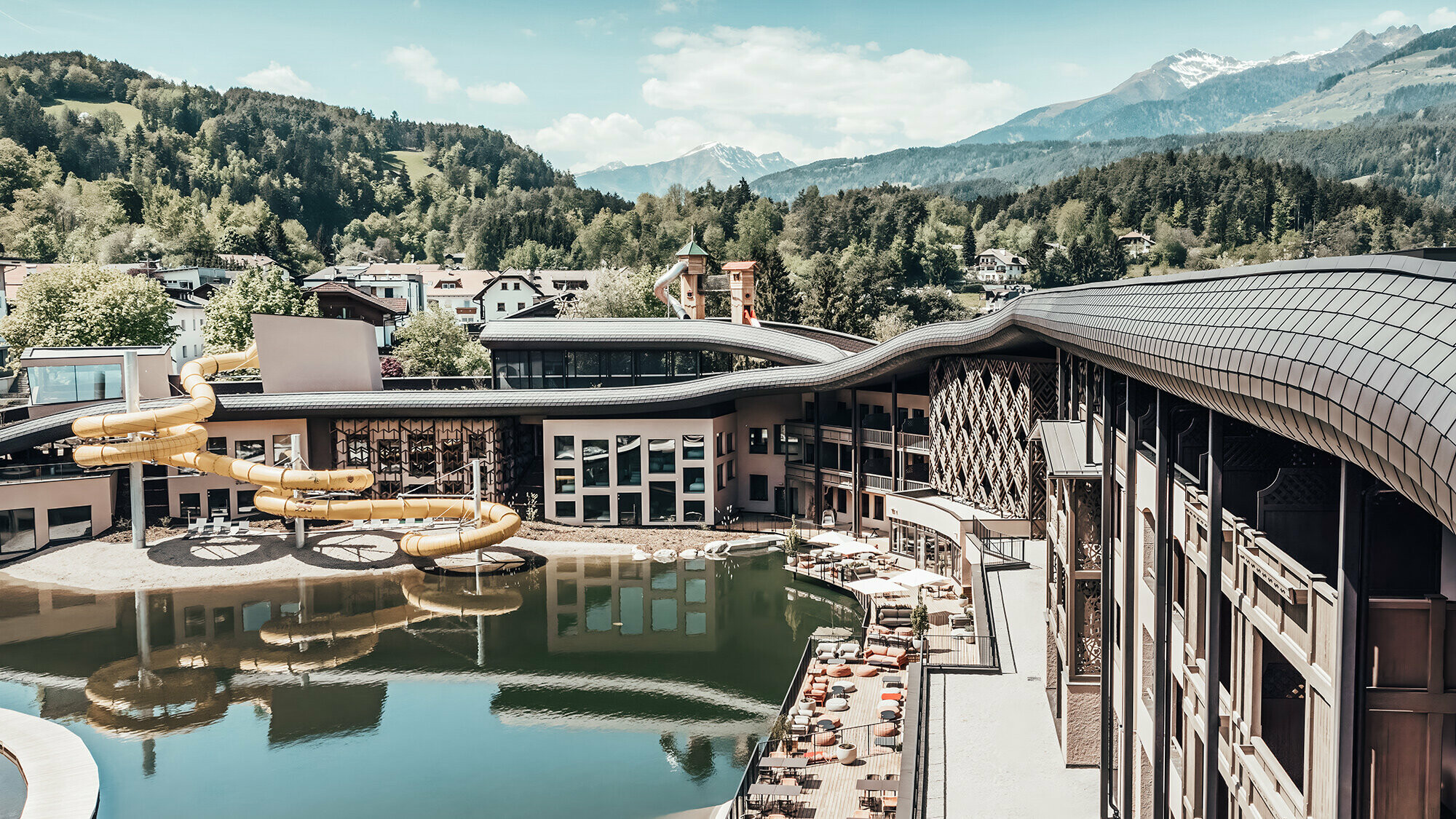 Überblick auf das Hotel mit See und Wasserrutsche. Im Hintergrund ist hügelige und bewaldete Fläche zu sehen.