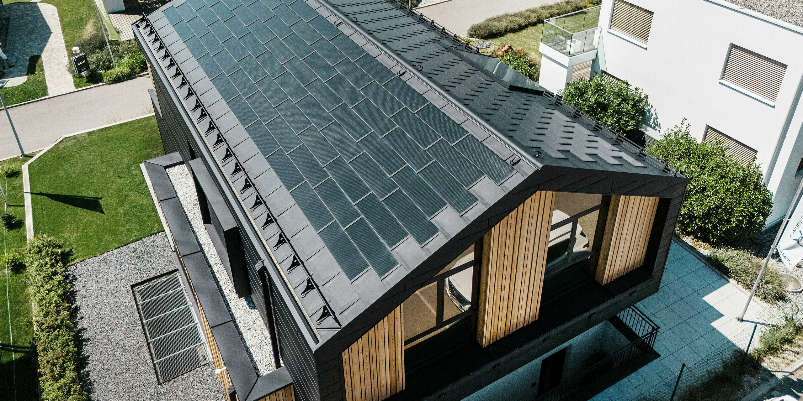 Vue aérienne de la maison témoin Autark dans le centre de maisons préfabriquées de Blaue Lagune, Vösendorf (AT), avec une toiture robuste en aluminium PREFA. La toiture est recouverte de tuiles solaires PREFA et de R.16 en P.10 noir. Le système photovoltaïque intégré garantit une production d'énergie durable en utilisant l'énergie du soleil. Le bardage en bois côté pignon complète l'aspect contemporain de la maison.