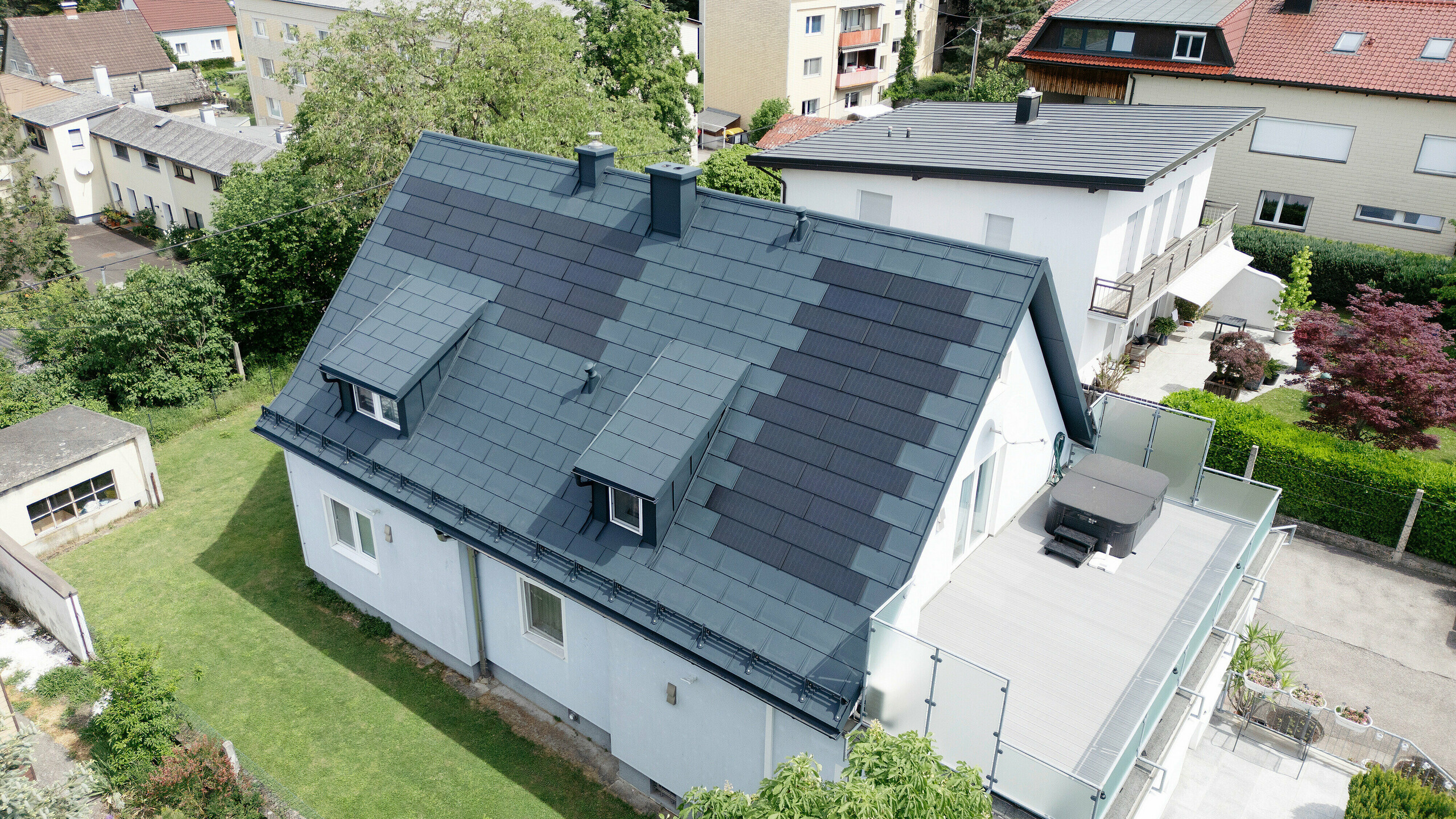 Vollständige Dachsanierung eines Einfamilienhauses in Leonding mit PREFA Solardachplatten in Anthrazit. Das Bild zeigt das Haus aus der Vogelperspektive nach der Sanierung. Die integrierten Solarmodule auf dem Dach bieten eine nachhaltige und energieeffiziente Lösung, die sowohl ästhetisch ansprechend als auch funktional ist. Die langlebigen und witterungsbeständigen PREFA Materialien tragen zu einer modernen und umweltfreundlichen Architektur bei, die den neuesten Standards für nachhaltiges Bauen entspricht.