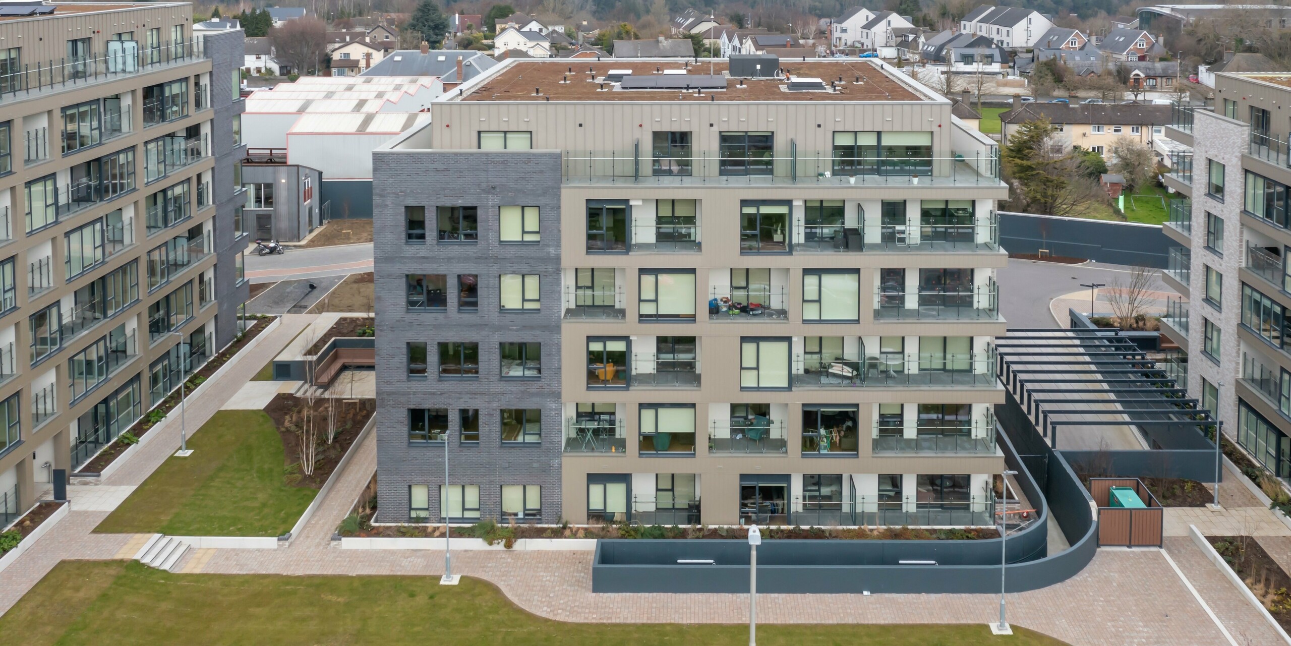 Drohnenaufnahme des Palmers Gate Wohnkomplexes in Palmerstown, Dublin, Irland. Der Gebäudekomplex umfasst mehrere Wohnblöcke mit bronzefarbenen PREFA Sidings und PREFALZ Fassadensystem. Die Fassade kombiniert hochwertige Blechverkleidungen mit Ziegelsteinelementen und großen Glasfenstern. Die Außenbereiche sind mit Grünflächen, Gehwegen und Pflanzkästen gestaltet, die den Bewohnern eine angenehme Umgebung bieten. Insgesamt wurden ca. 1.700 m² PREFA Sidings und ca. 1.200 m² PREFALZ Fassadensystem verwendet. Die Aluminiumprodukte sind für ihre Langlebigkeit und ästhetische Optik bekannt.