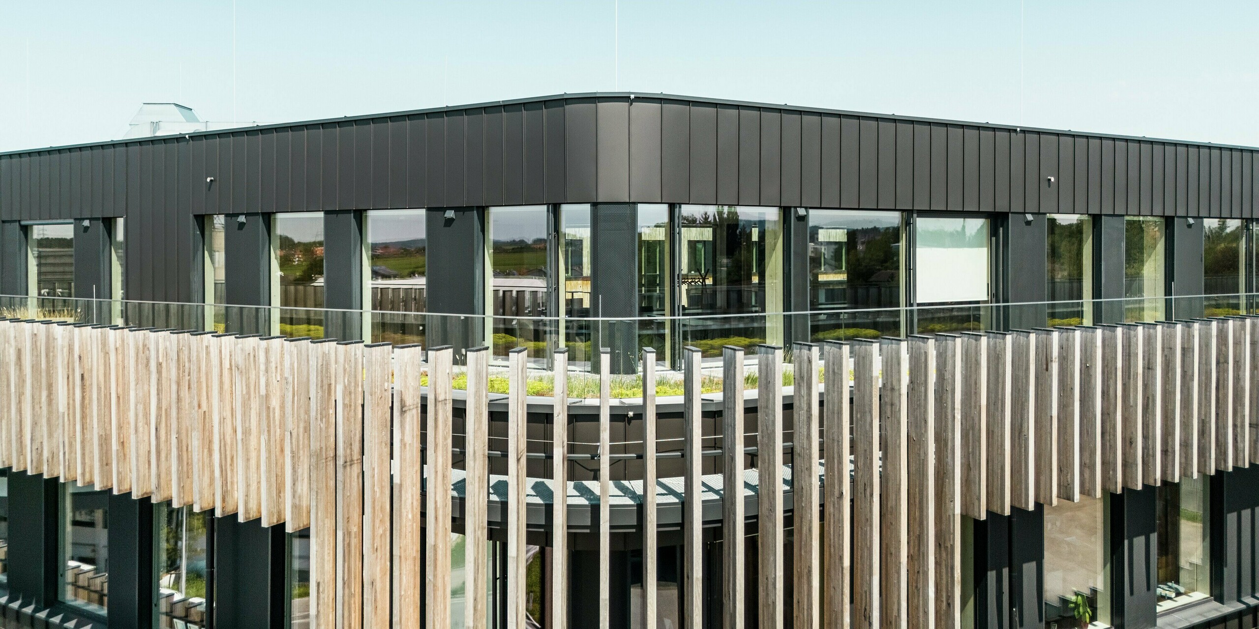 Le Team 7 World à Ried im Innkreis, en Haute-Autriche, présente une combinaison impressionnante de façade PREFALZ dans la couleur spéciale noir-brun et d'éléments en bois verticaux. Le revêtement en aluminium de haute qualité allie durabilité, résistance aux intempéries et design moderne. Associé aux grands vitrages, cela crée une interaction harmonieuse entre fonctionnalité, esthétique et durabilité. Le ciel bleu vif s'élève au-dessus de l'attique.