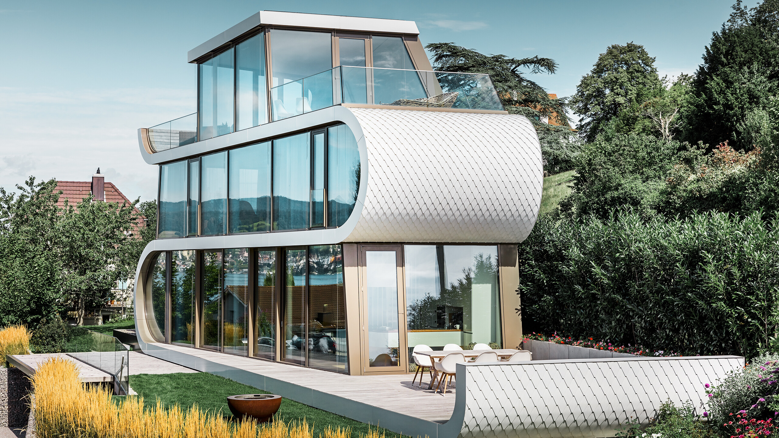 Aufnahme des modernen Flexhouses am Zürichsee; Mit seiner außergewöhnlichen Gebäudeform fällt das Flexhouse immer auf. Die Geschoßtrennungen ziehen sich als weißes Band geschwungen durch das Gebäude. die runden Fassadenelemente wurden mit den Alu-Schindeln von PREFA in Reinweiß verkleidet. 