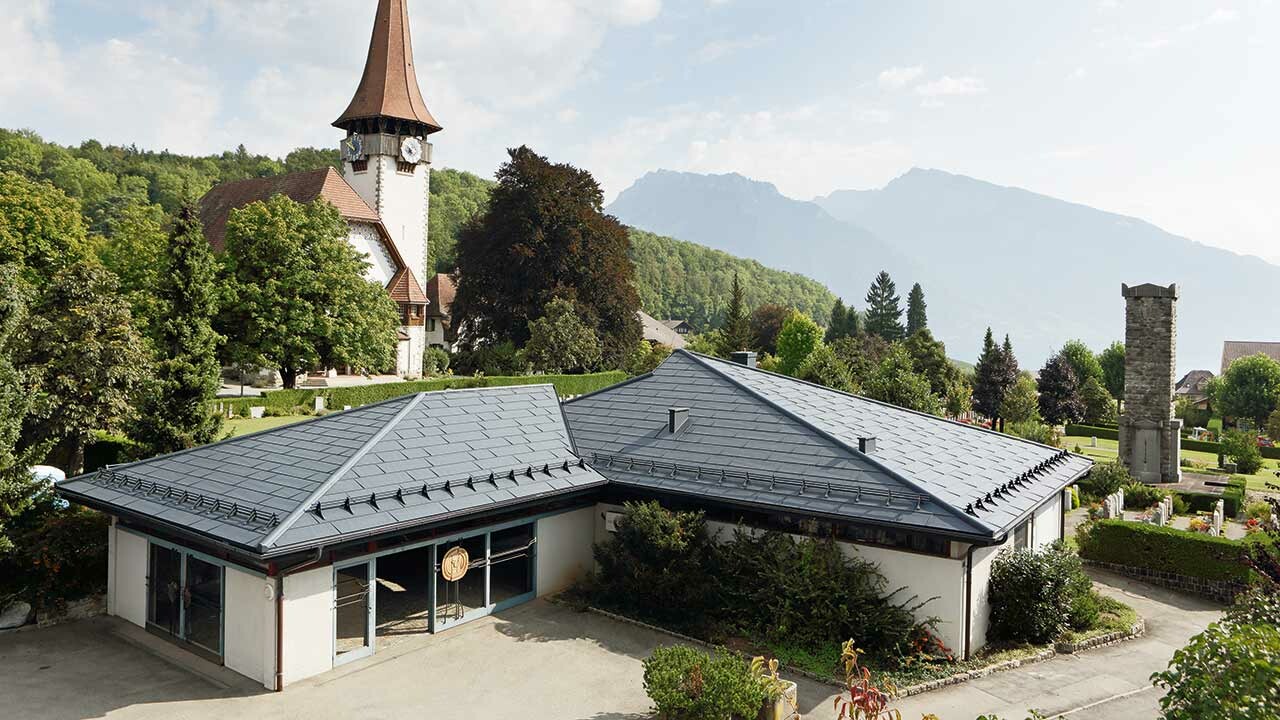 Aufbahrungshalle mit FX.12 Dachpaneelen von PREFA in Anthrazit, im Hintergrund steht eine Kirche und daneben befindet sich der Friedhof.