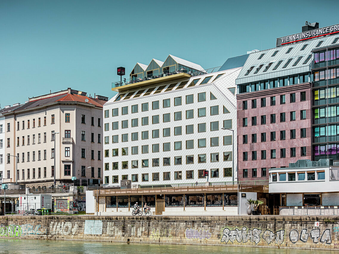 Veduta delle rive del canale del Danubio a Vienna con una fila di edifici di diversa architettura. In primo piano l'hotel "THE ROCK Radisson RED Vienna", riconoscibile per la caratteristica facciata in alluminio PREFA bianco con tetto 44 × 44 e diamanti a parete in P.10 bianco puro.