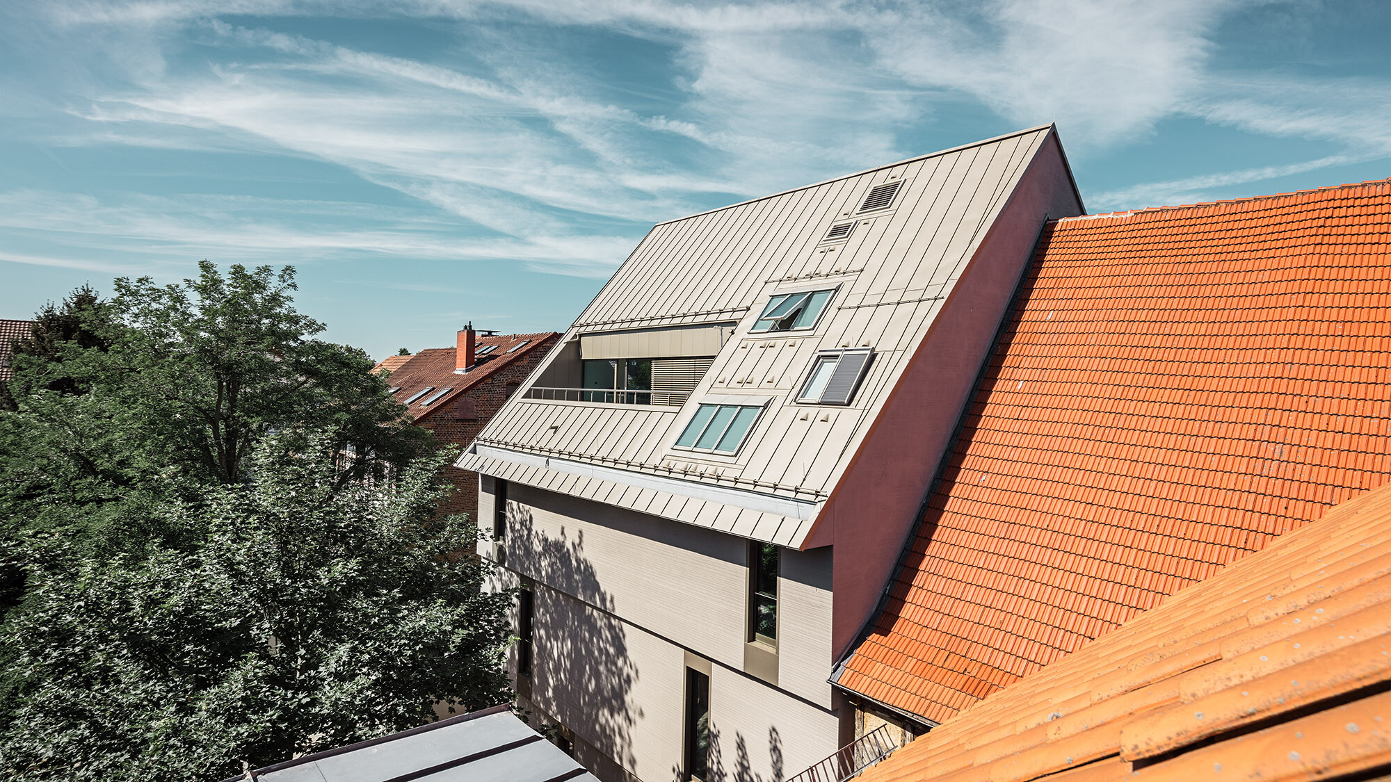 Une vue latérale de ce toit exceptionnel : à gauche et à droite se trouvent des bâtiments recouverts de tuiles plates de couleur rouille, devant lesquels se dressent des arbres.