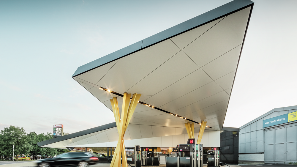 [Translate to Italiano:] Seitliche Ansicht der Tankstelle mit einer Untersicht des Daches, welches auf zwei goldenen Stützengruppen aufliegt.