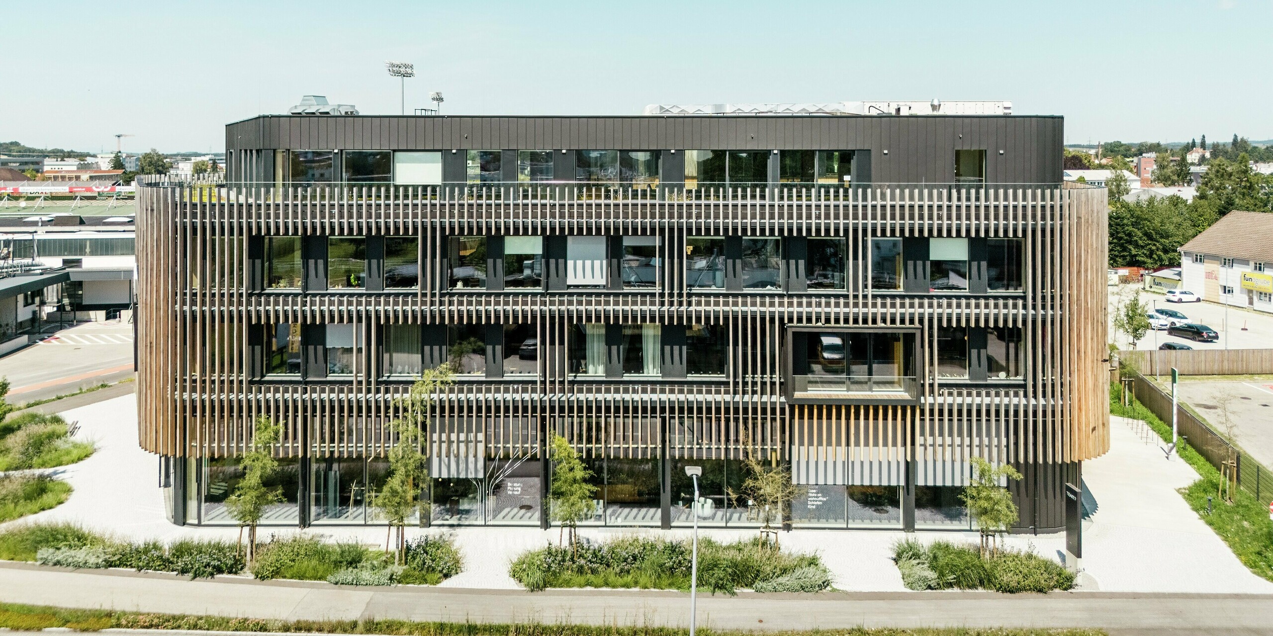 Vue latérale du complexe immobilier de Team 7 Welt à Ried im Innkreis. Les bâtiments combinent une façade PREFALZ noir-brun avec d'élégants éléments verticaux en bois et de grandes surfaces vitrées. Le revêtement innovant en aluminium offre durabilité, résistance aux intempéries et une esthétique moderne. Avec ses lignes claires, la façade s'harmonise parfaitement avec le paysage environnant et souligne les exigences de l'architecture contemporaine.