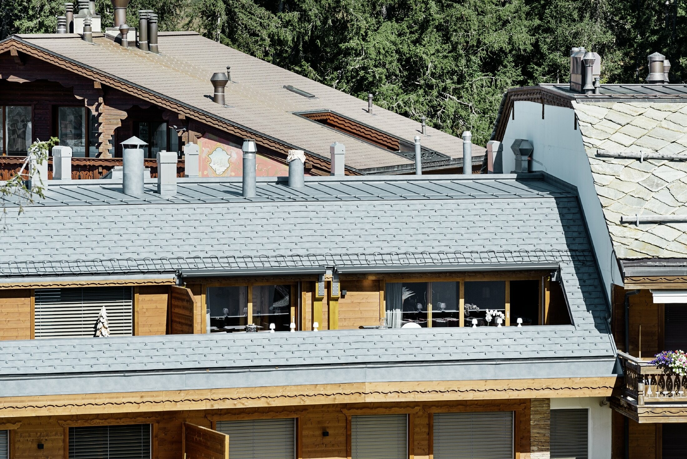 Casa plurifamiliare in Crans-Montana con montagne sullo sfondo e una facciata con elementi decorativi in legno e con una copertura in scandole PREFA grigio pietra