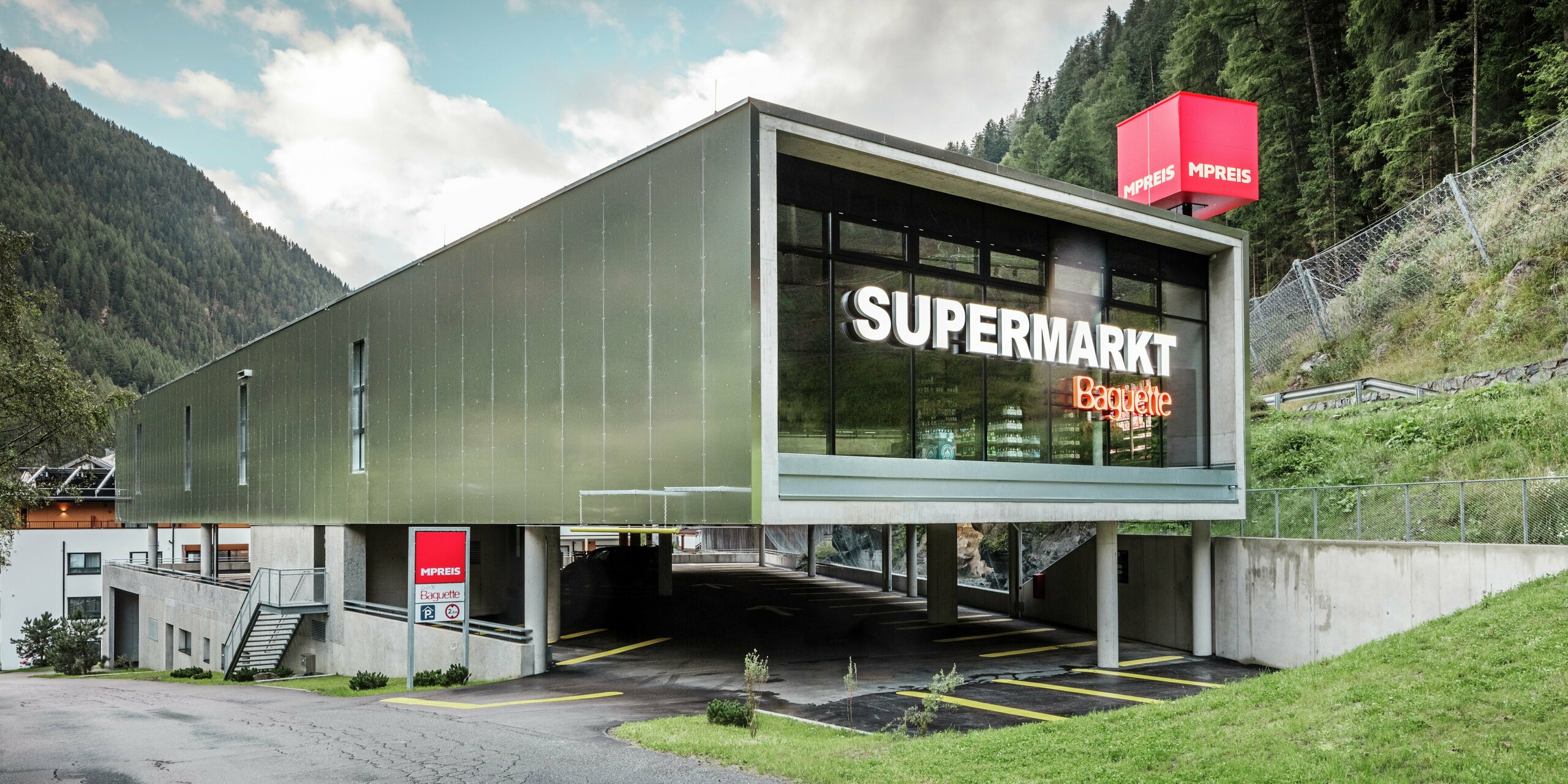 Zufahrt des MPREIS-Supermarktes in Sölden, Tirol, mit Aluminium Verbundplatten in der Sonderfarbe Reflective Natural. Die moderne Fassade spiegelt die umliegende Bergwelt wider. Großzügige Glasfronten, ein aufgeständertes Parkdeck und klare Linien verbinden Funktionalität mit zeitgemäßem Design im alpinen Raum.