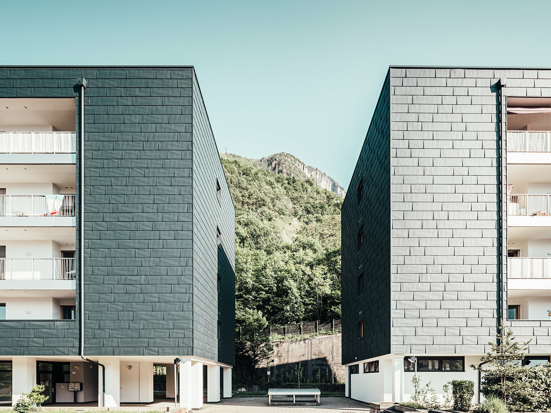 Prise de vue des deux côtés de l'immeuble aux façade à la teinte anthracite PREFA. Entre les deux, en arrière-plan, on aperçoit la montage et ses arbres.