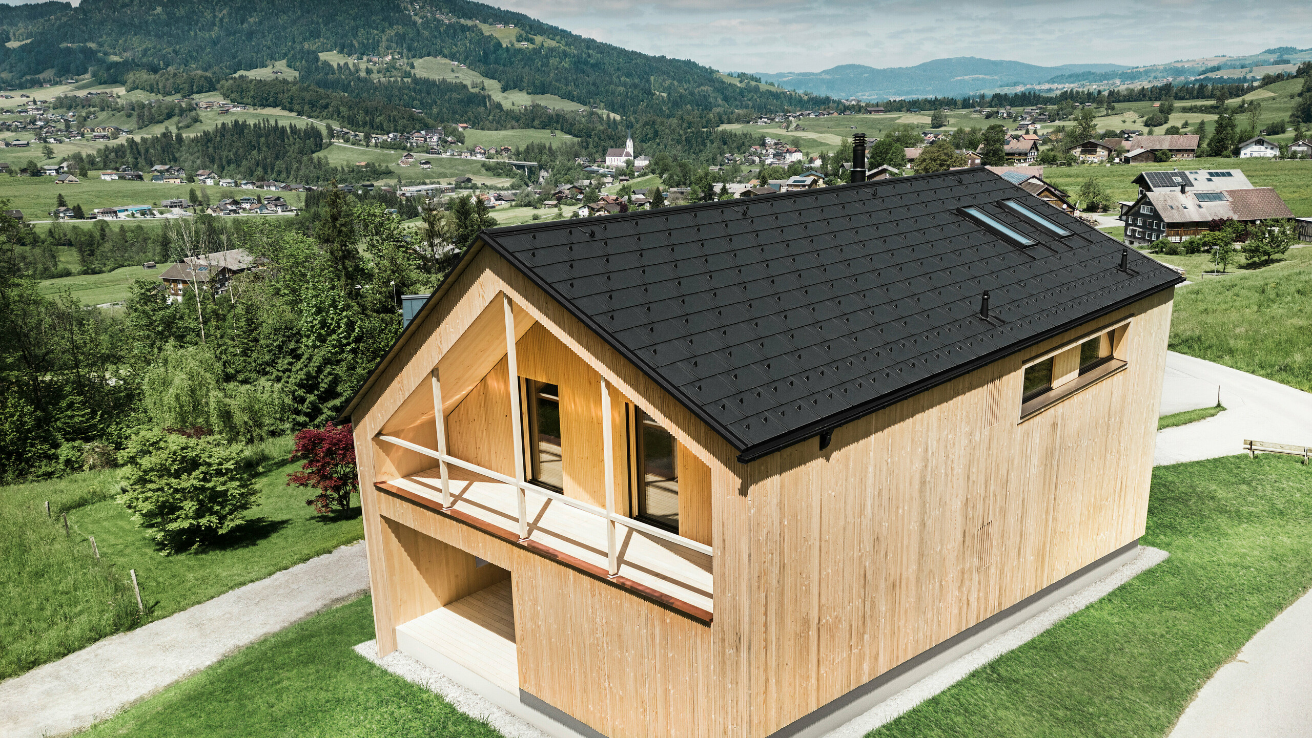 Dieses Foto zeigt ein ökologisches Vorzeigehaus in Egg, das sich mit seiner PREFA Solardachplatte und den schwarzen Dachplatten R.16 gegen die malerische Kulisse eines österreichischen Dorfes abhebt. Die Solarelemente sind nahtlos in die Dachstruktur eingebettet und liefern Energie aus einer erneuerbaren Energiequelle, während das natürliche Holz der Fassade und die sattgrüne Umgebung eine Verbindung von Nachhaltigkeit und Lokalität darstellen. Der Blick auf die umliegende dörfliche Struktur mit traditionellen Bauten im Hintergrund verleiht dem Bild eine zusätzliche Dimension und betont die gelungene Integration moderner, umweltfreundlicher Bauweisen in gewachsene Landschaften.