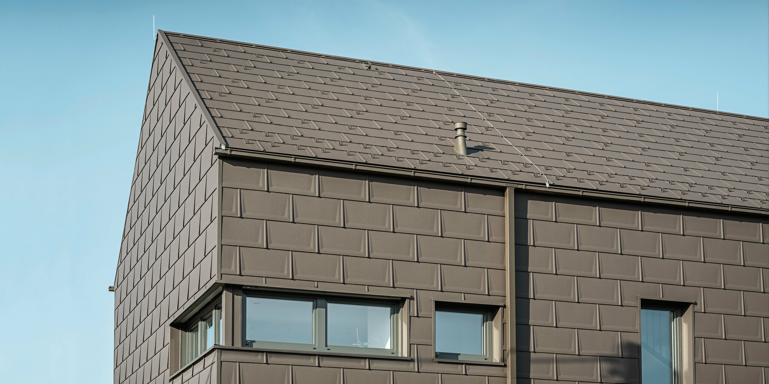Detailaufnahme eines Einfamilienhauses in Neukirchen, Österreich, mit einer Dach- und Fassadenverkleidung aus PREFA R.16 Dachplatten in P.10 braun. Die präzise Überlappung der Platten sorgt für ein einheitliches und ansprechendes Erscheinungsbild, das moderne Ästhetik mit Funktionalität verbindet. Die integrierte PREFA Dachentwässerung in Form einer Kastenrinne und eines Vierkantrohrs in P.10 Braun zeigt das Engagement von PREFA für hochwertige und optisch ansprechende Bauelemente.