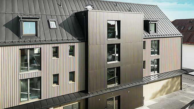 La photo montre un bâtiment moderne à Feldkirchen, conçu par l'architecte Stefan Heigl. La façade est recouverte de Prefalz de couleur bronze, tandis que le toit à pignon est recouvert de Prefalz de couleur gris noir. Les grandes fenêtres et lucarnes apportent beaucoup de lumière naturelle à l’intérieur du bâtiment. La conception allie des matériaux de haute qualité et une architecture élégante, à la fois fonctionnelle et esthétique. Cette méthode de construction moderne et l'utilisation de matériaux Prefalz confèrent au bâtiment un aspect contemporain et élégant.