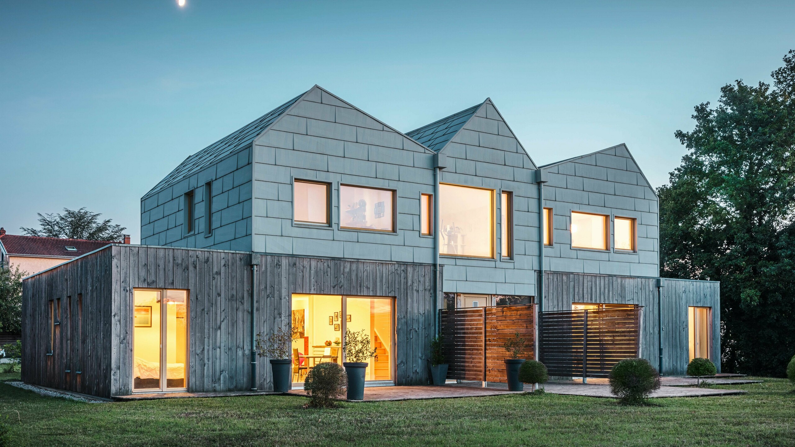 Doppelhaus bei Abenddämmerung mit leuchtenden Innenräumen, die durch große Fenster sichtbar sind. Die Aluminiumpaneele von PREFA in FX.12-Optik fügen sich nahtlos in das moderne Design ein. Das Gebäude ist von einem gepflegten Garten umgeben.