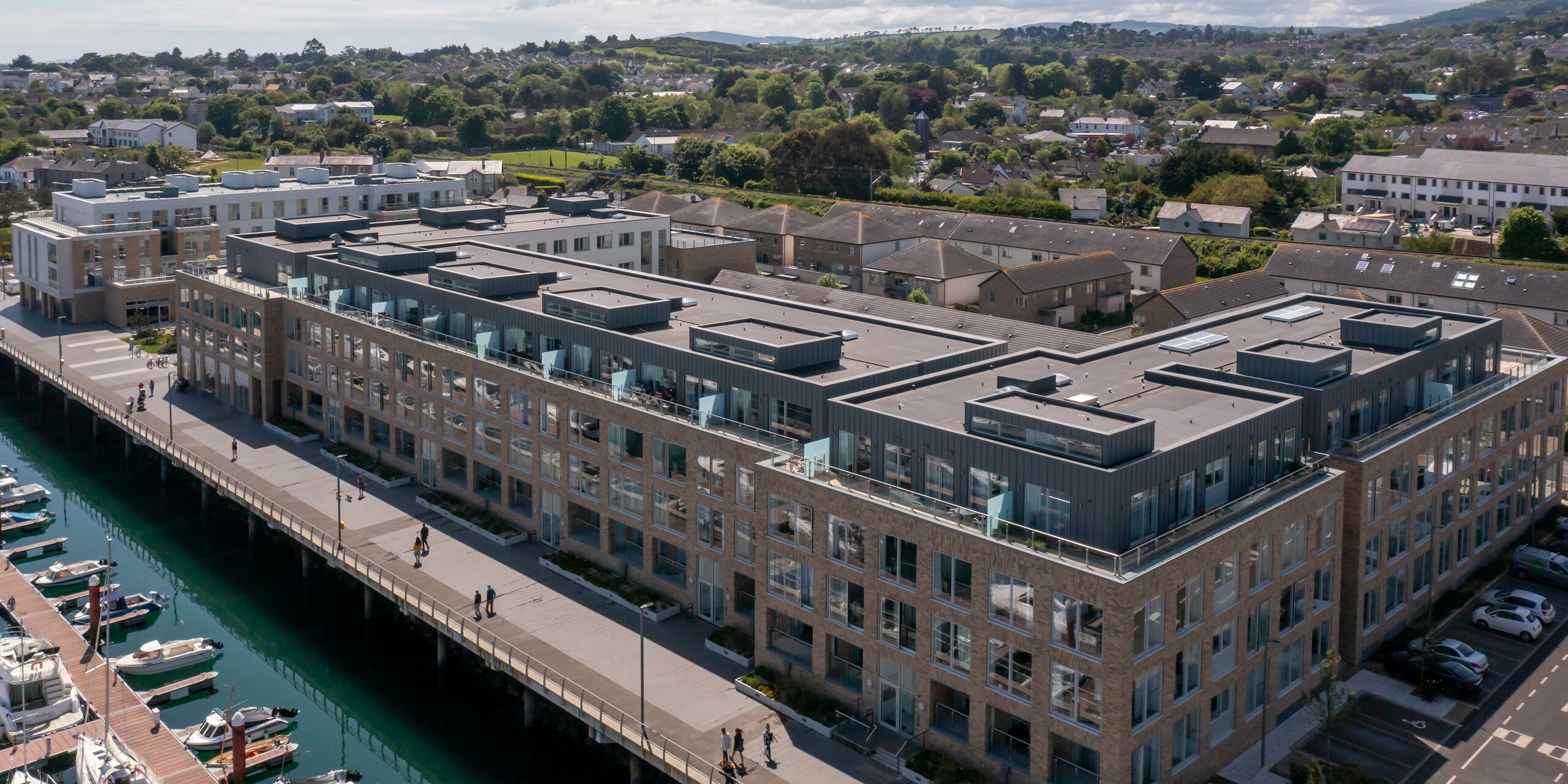 Weitwinkel-Vogelperspektive auf Marina Village Greystones in Wicklow, Irland – Moderne Wohnsiedlung am Wasser mit direkter Lage am Yachthafen. Der Gebäudekomplex ist mit robustem PREFALZ in den Farben P.10 Prefaweiß und P.10 Hellgrau verkleidet. Die elegante Architektur umfasst große Glasfronten, geräumige Balkone und eine hochwertige Fassadengestaltung aus Blech. Die Verwendung von PREFALZ-Aluminium bietet dauerhaften Schutz und eine ansprechende Optik. Umgeben von malerischer Landschaft steht das Wohnprojekt für exklusives Wohnen am Meer.