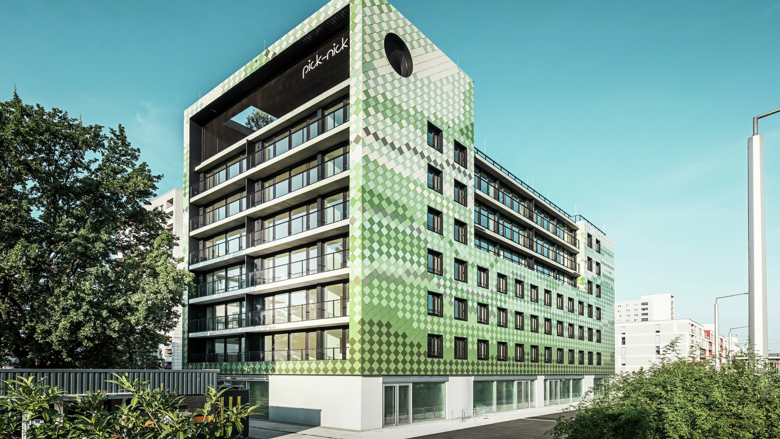 Bâtiment résidentiel et commercial « Pick-Nick » à Dresde avec une façade moderne en panneaux d'aluminium PREFA dans différents tons verts et beiges. La façade distinctive du bâtiment combine des lignes claires avec un jeu de couleurs dynamique et est en outre enrichie de grandes surfaces vitrées. Les accents architecturaux comprennent l'inscription « Pick-Nick » sur le bord supérieur du bâtiment et le jardin sur le toit vert.