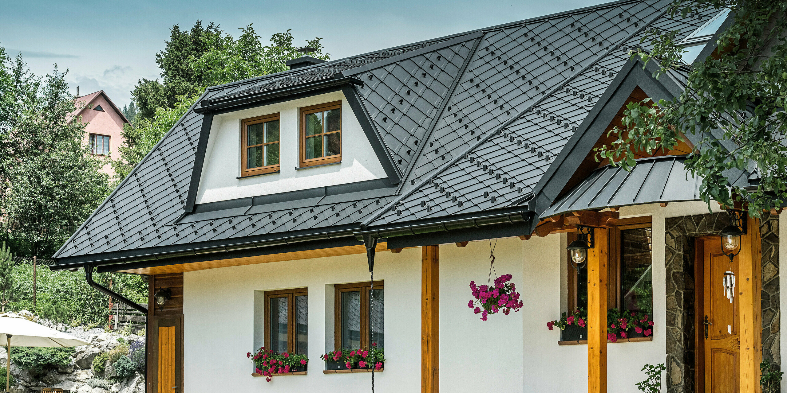 Vue de face d'une maison familiale traditionnelle à Stara Bystrica, en Slovaquie, avec un toit en losanges PREFA 29 × 29 en P.10 noir. Le toit au look mémorable comprend des lucarnes et des auvents trapézoïdaux. Les lignes claires et les fenêtres soigneusement placées confèrent au bâtiment une apparence attrayante. Des compositions florales et un aménagement de jardin bien entretenu complètent le tableau général.