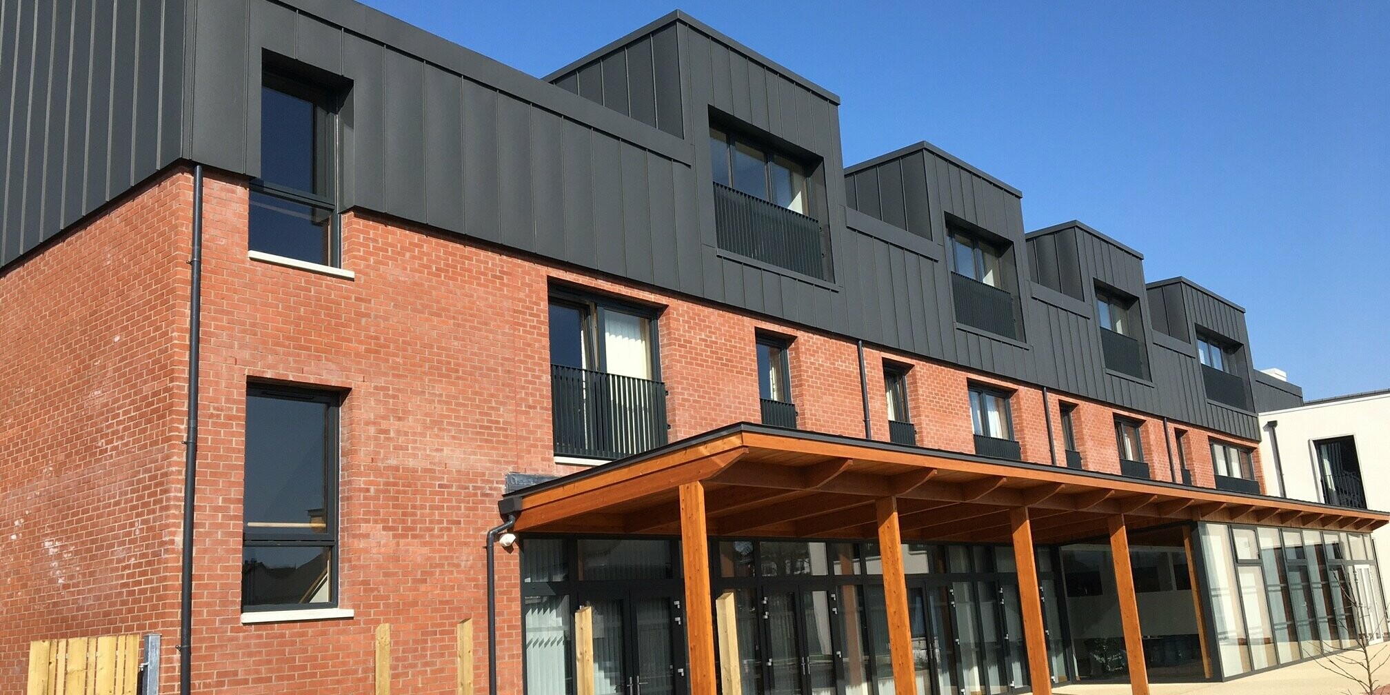 Vue depuis le jardin de Harberton Hall à Belfast, en Irlande du Nord. La toiture à joint debout en aluminium PREFALZ gris sombre rencontre la façade en briques. Un espace extérieur vert et des lucarnes à toit plat renforcent la combinaison harmonieuse de l'architecture moderne et du style traditionnel dans ce complexe de vie assistée.