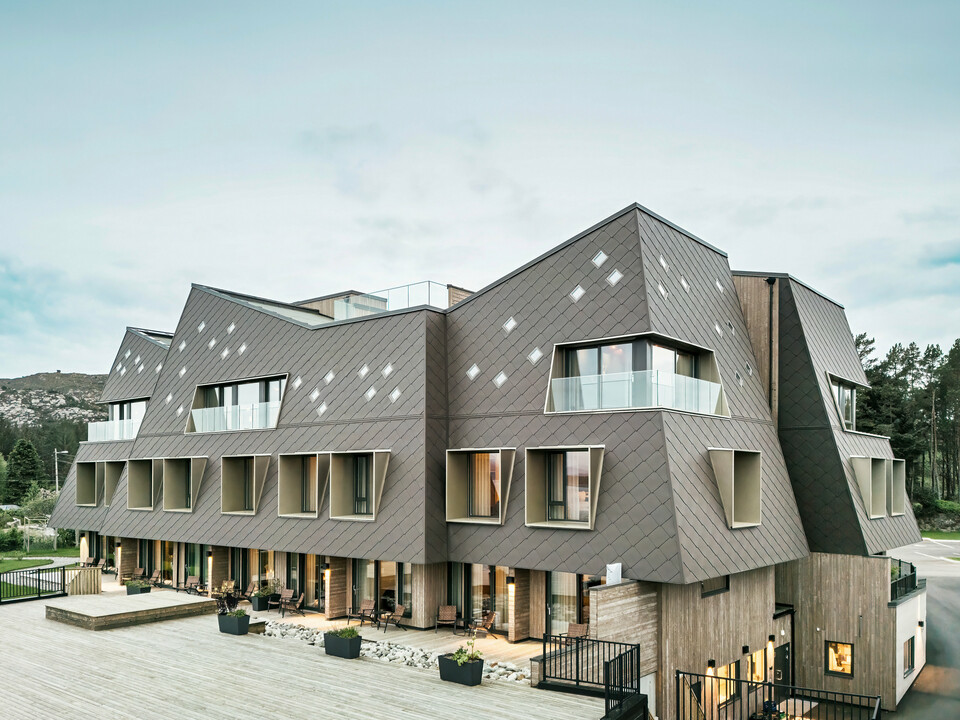 Blick auf die Terrasse des Hotels Beckerwyc House in Bekkjarvik, Norwegen. Das moderne Gebäude wird von einer einer markanten Aluminiumfassade aus PREFA Wandrauten 44 × 44 in Nussbraun geschmückt. Die futuristische Architektur mit schrägen Dachflächen und hervortretenden Fensterelementen wird durch die großzügigen Außenbereiche ergänzt. Die natürliche Farbgebung der Fassade passt sich harmonisch in die umgebende Landschaft ein und unterstreicht das zeitgemäße Design des Gebäudes.