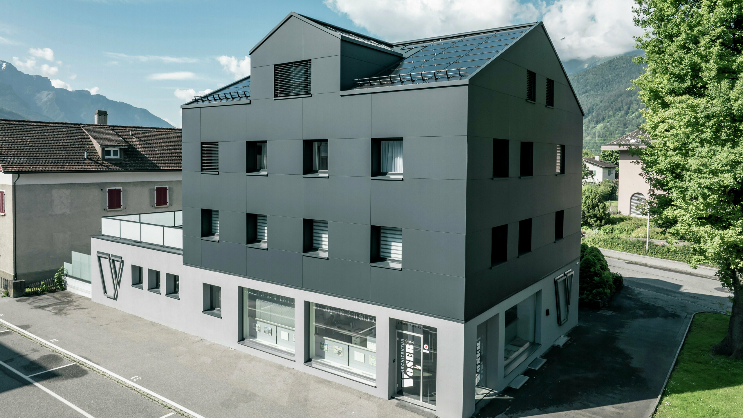 Modernisiertes Wohn- und Geschäftsgebäude nach der Fassadensanierung mit PREFABOND Aluminium Verbundplatten in Anthrazit. Die klare, elegante Linienführung der neuen Fassade verleiht dem Gebäude ein modernes Erscheinungsbild. Im Erdgeschoss befindet sich das Architekturbüro Voser. Die Umgebung mit Bäumen und Bergen rundet die harmonische Optik ab.