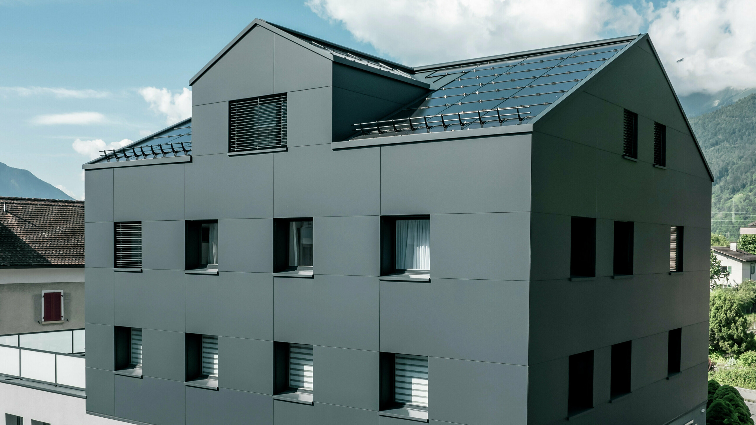 Modernisiertes Wohn- und Geschäftsgebäude nach der Fassadensanierung mit PREFABOND Aluminium Verbundplatten in Anthrazit. Die klare, elegante Linienführung der neuen Fassade verleiht dem Gebäude ein modernes Erscheinungsbild. Im Erdgeschoss befindet sich das Architekturbüro Voser. Die Umgebung mit Bäumen und Bergen rundet die harmonische Optik ab.