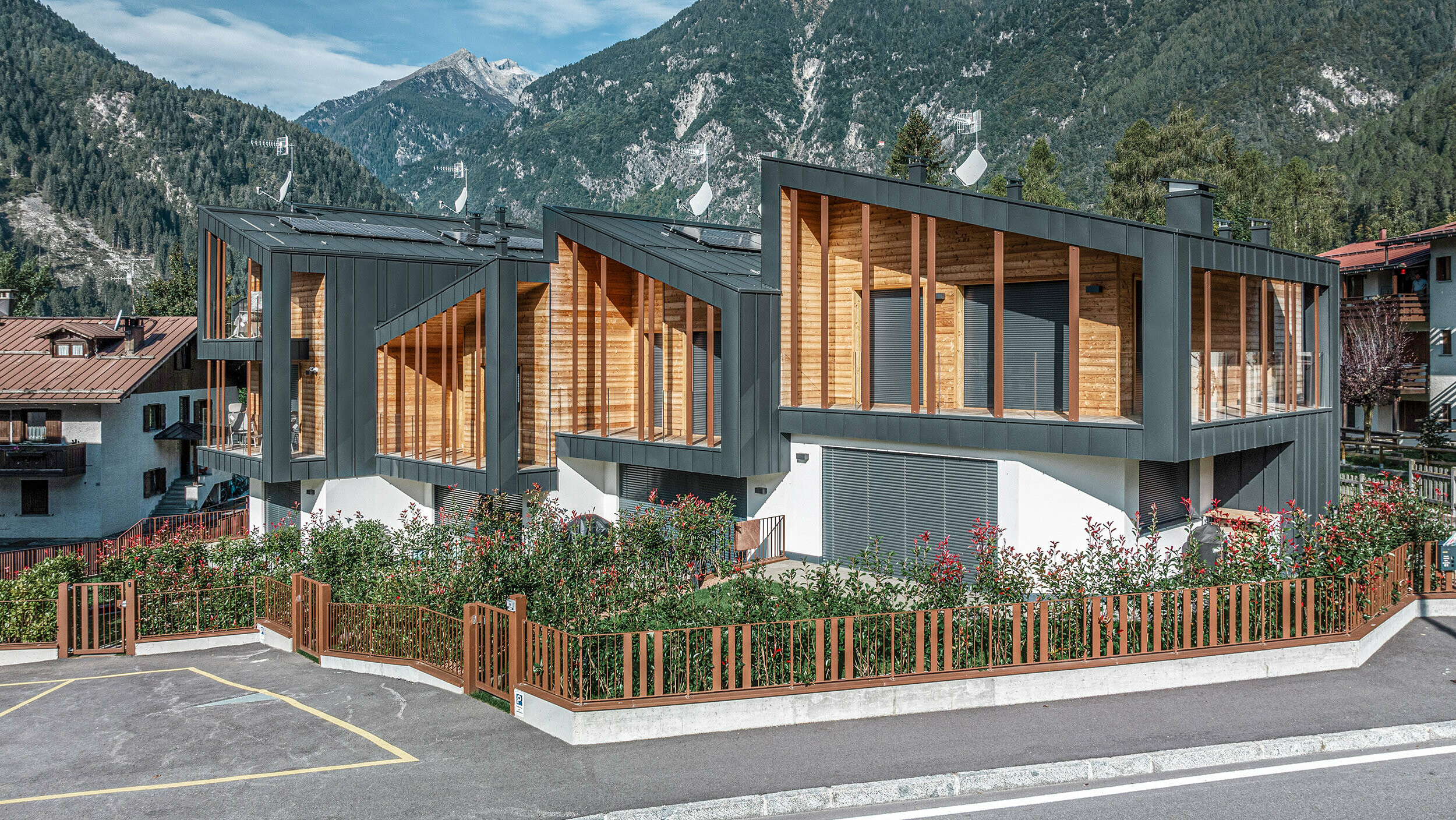 Nouvelle construction de chalets modernes avec toits monopentes en métal Prefalz, façades associant métal gris foncé et éléments en bois, le tout dans un paysage de montagne et sous un ciel dégagé. Les larges fenêtres et terrasses sont complétées par des espaces verts et un parking entouré d’une clôture.