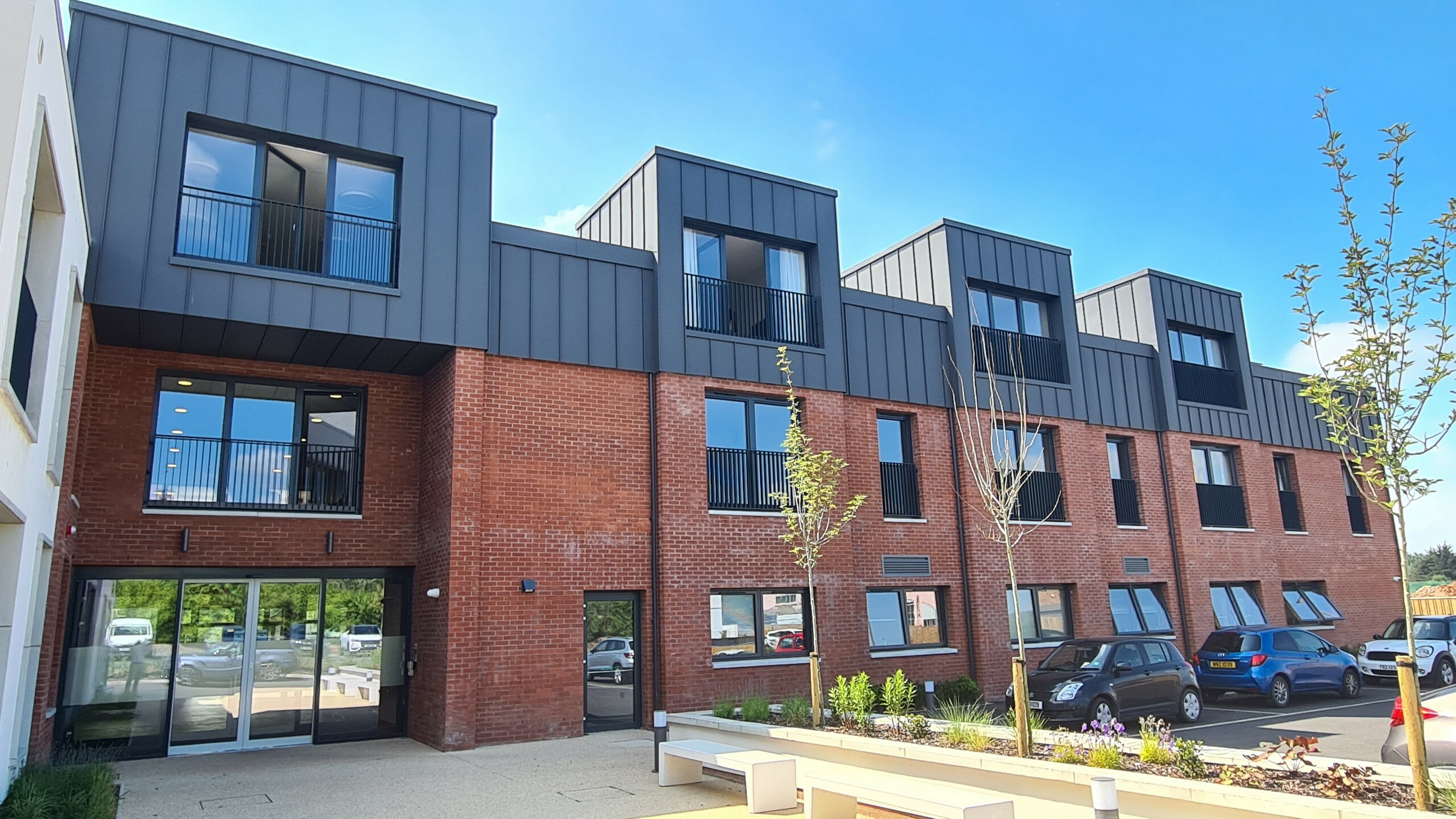 Vue de face de Harberton Hall à Belfast, en Irlande du Nord. La toiture à joint debout en PREFALZ P.10 gris sombre complète parfaitement la façade en briques. De grandes fenêtres et des lucarnes à toit plat soulignent la conception innovante du bâtiment de vie assistée et créent une atmosphère accueillante. La porte d'entrée vitrée donne un aperçu de l'intérieur de l'établissement.