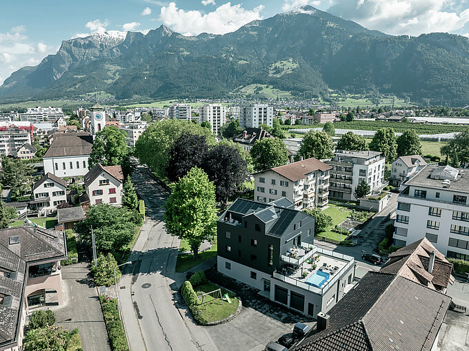 Vogelperspektive auf ein modernes Geschäftsgebäude mit einer sanierten Fassade aus PREFABOND Aluminium-Verbundplatten in Anthrazit. Das Gebäude fügt sich harmonisch in die grüne Umgebung und die umliegenden Wohnhäuser ein, während im Hintergrund eine beeindruckende Berglandschaft zu sehen ist.