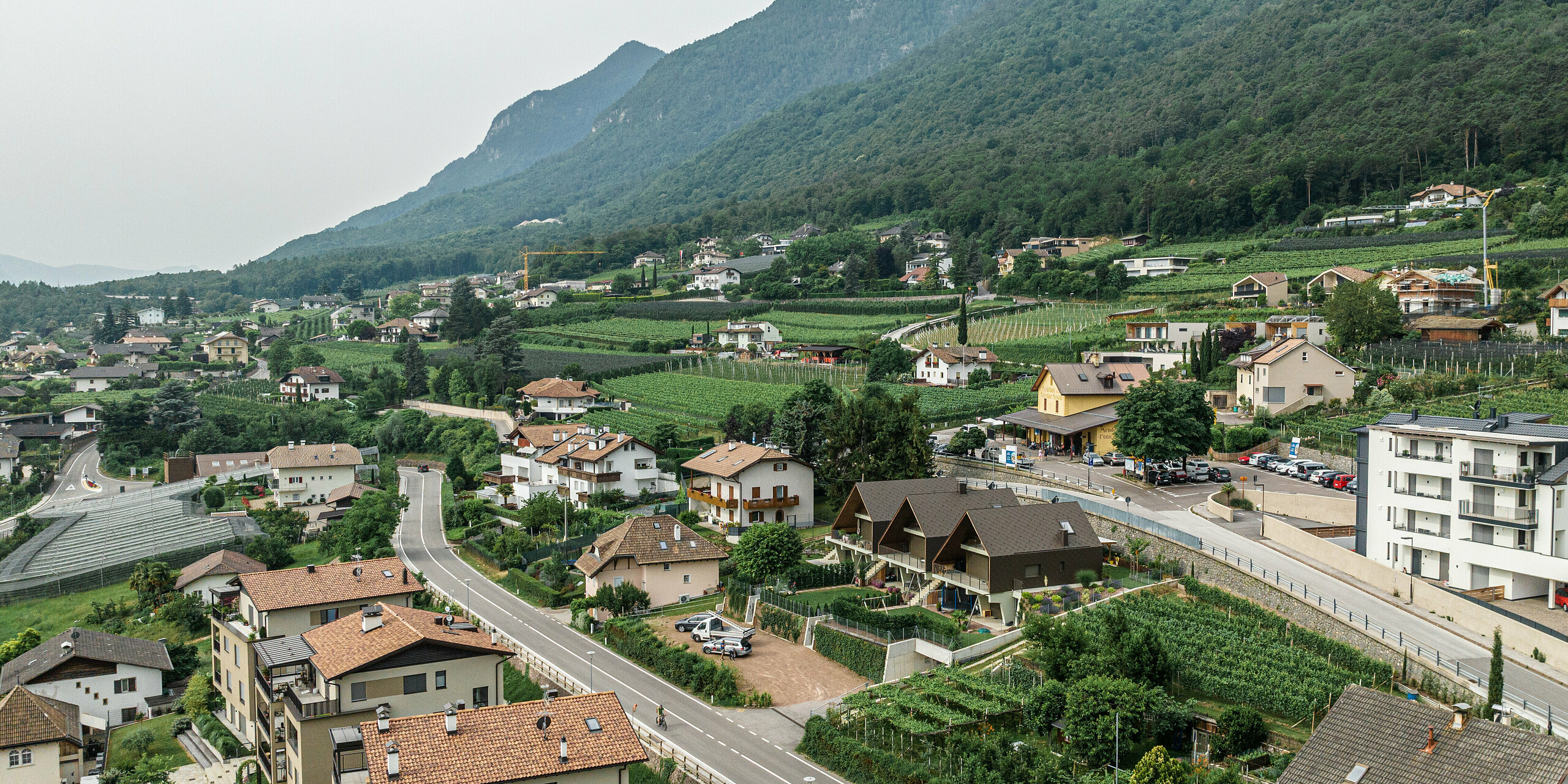 Drohnenaufnahme eines Dorfes in einem bergigen Gebiet in Südtirol. Im Zentrum des Bildes stehen drei Reihenhäuser, hervorgehoben durch eine markante Aluminiumhülle aus PREFA Dach- und Wandrauten 44x44 in der Farbe P.10 Braun. Die moderne Architektur fügt sich nahtlos in die traditionelle Umgebung ein, umgeben von üppigen grünen Weinbergen und dichten Wäldern.